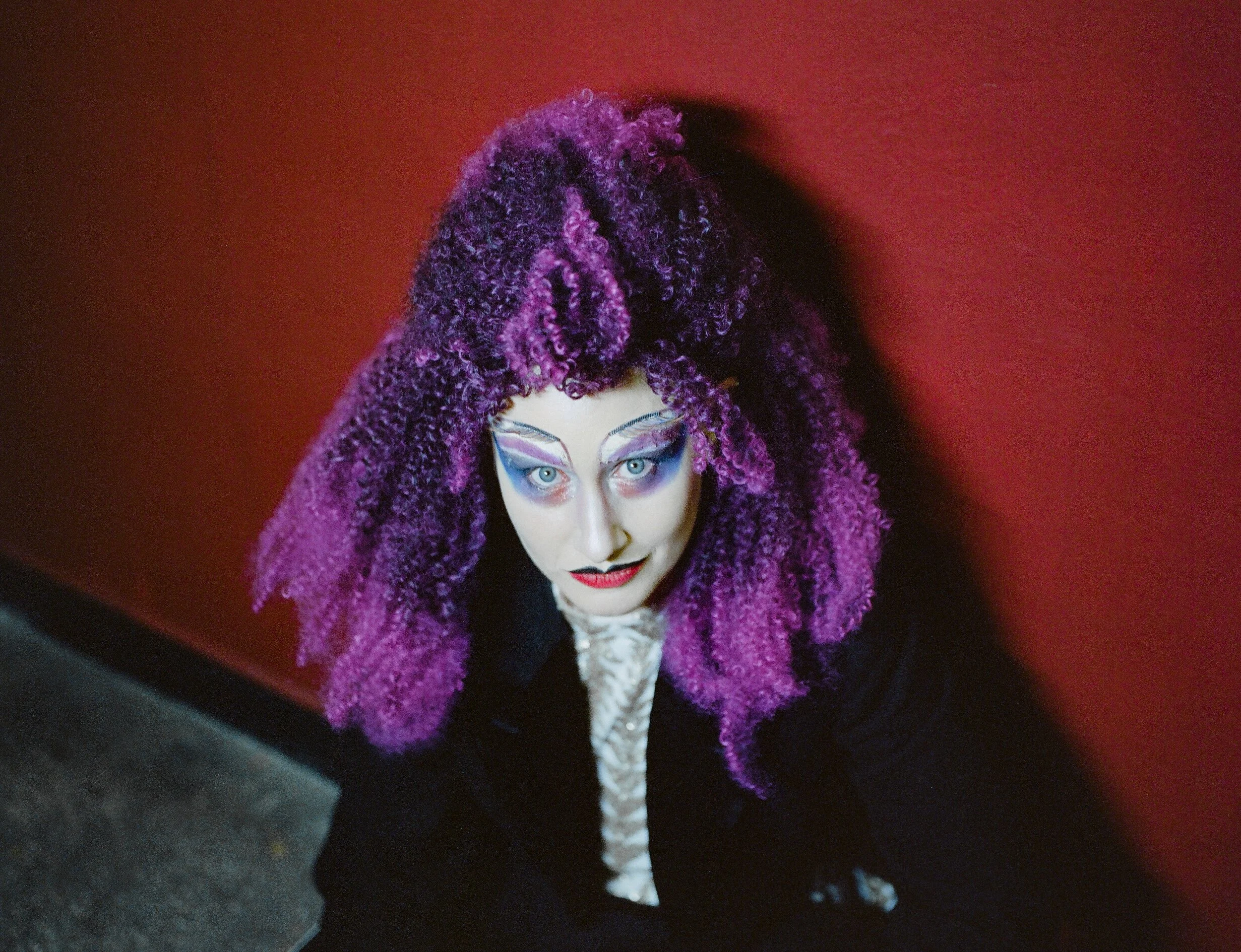 A woman with colorful eye makeup and violet curly hair, wearing a purple jacket and black clothing, looking up in a setting with a red background.