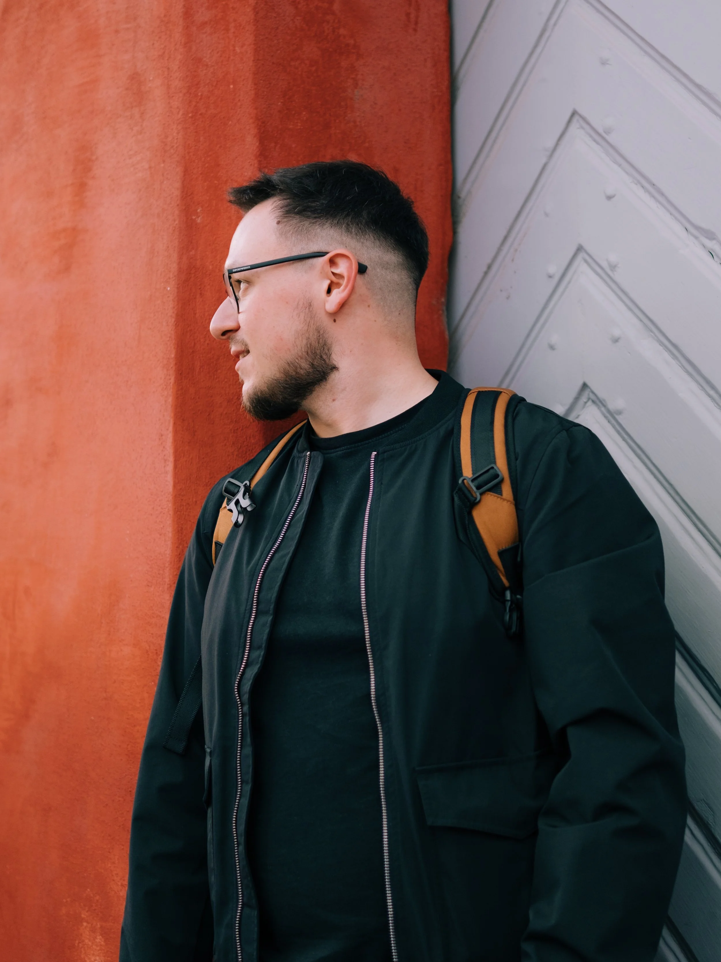 A man with short dark hair, glasses, and facial hair, wearing a black jacket and an orange backpack, is leaning against a wall with red and white panels.