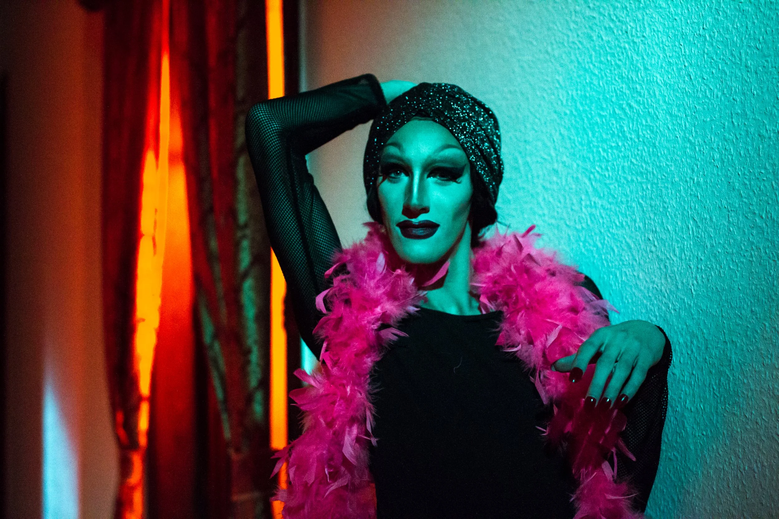 A person with dramatic makeup and a sparkly headwrap, wearing a pink feather boa, posed against a textured wall illuminated by colorful neon lights in red, orange, teal, and purple hues.