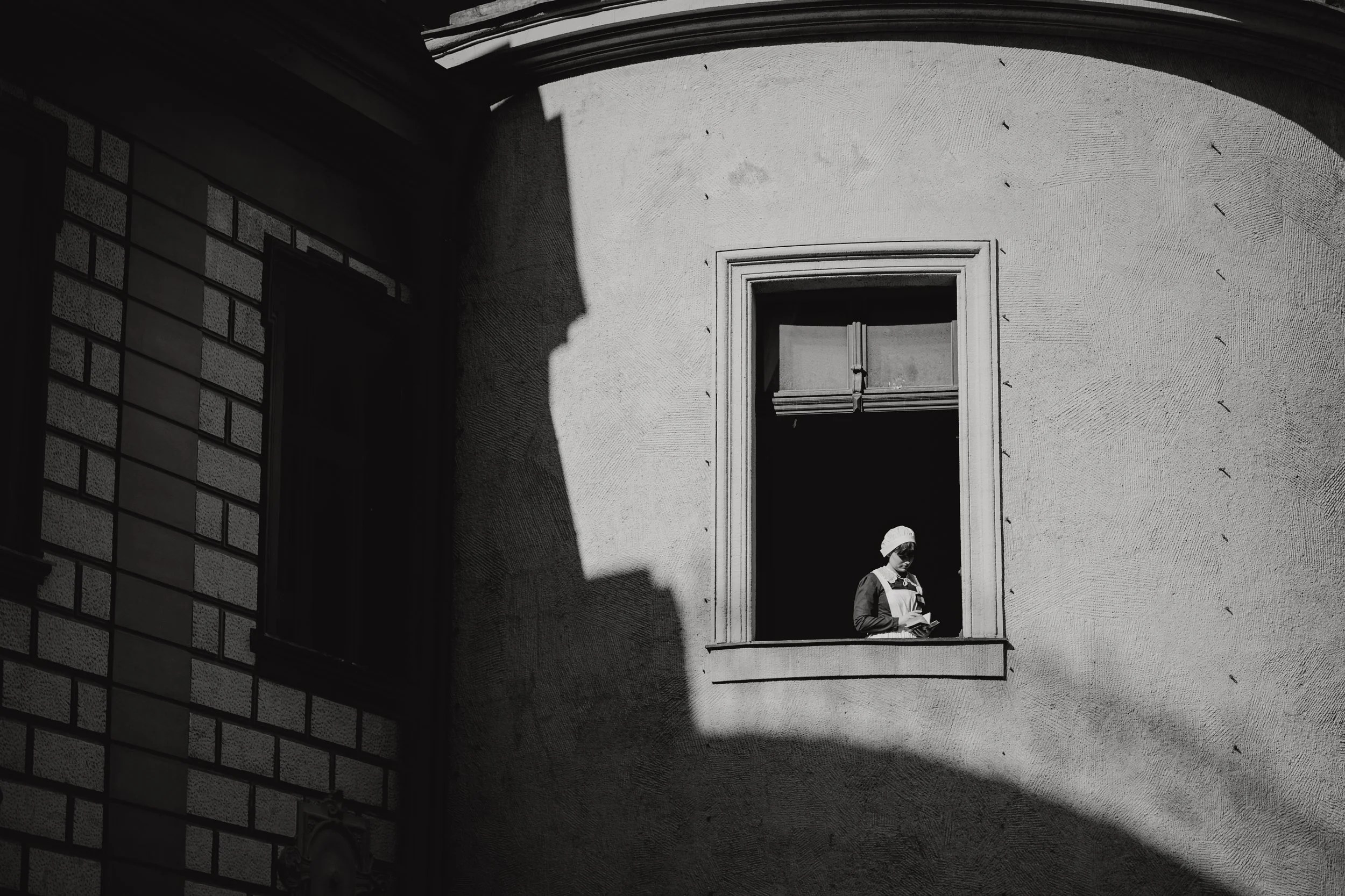 A woman wearing a headscarf and dress standing in a window of a building, with sunlight casting shadows on the adjacent wall.