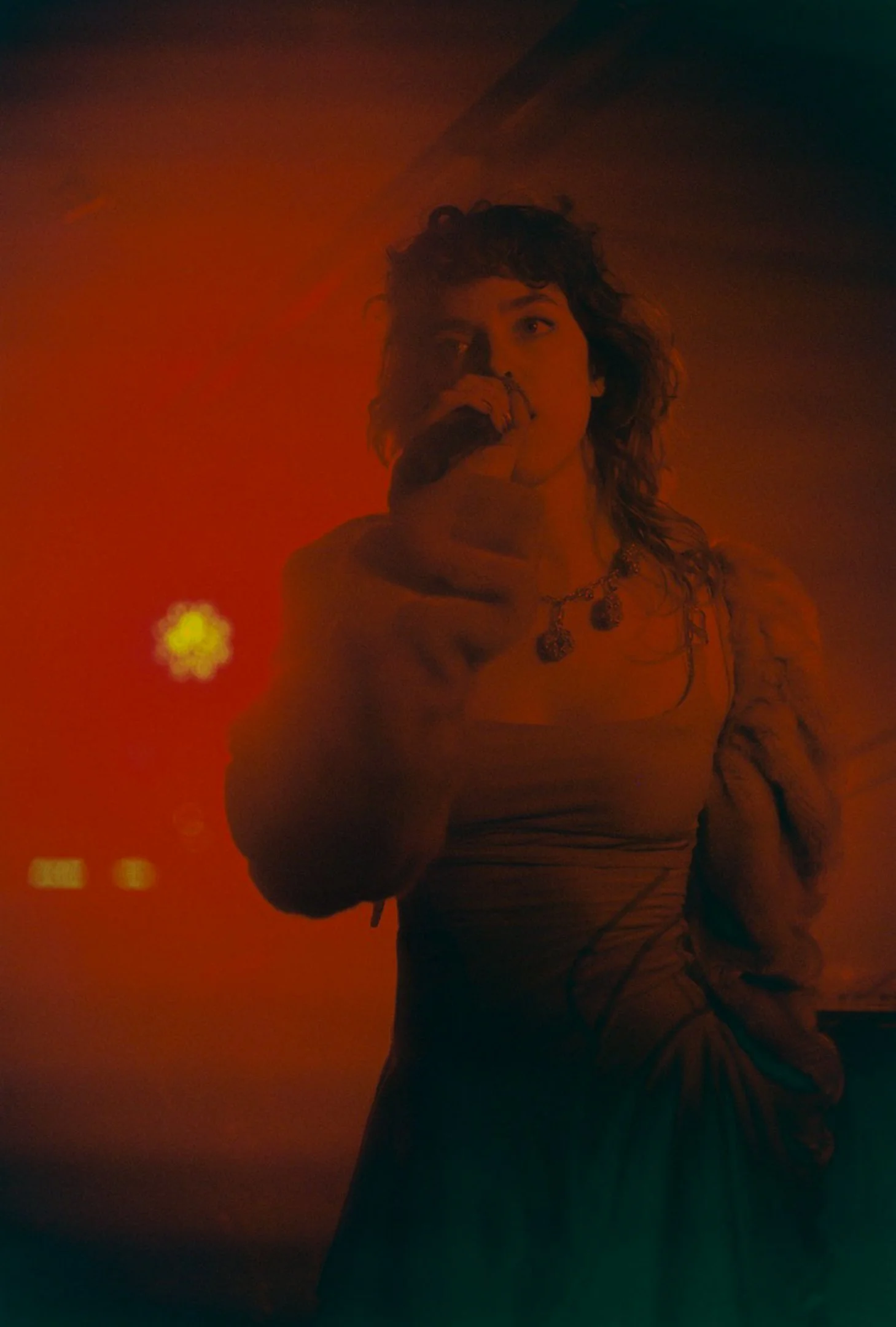 Une femme avec des cheveux bouclés et longs, portant un vêtement moulant et un collier, chante dans une ambiance sombre avec un éclairage rouge, en tenant un microphone.