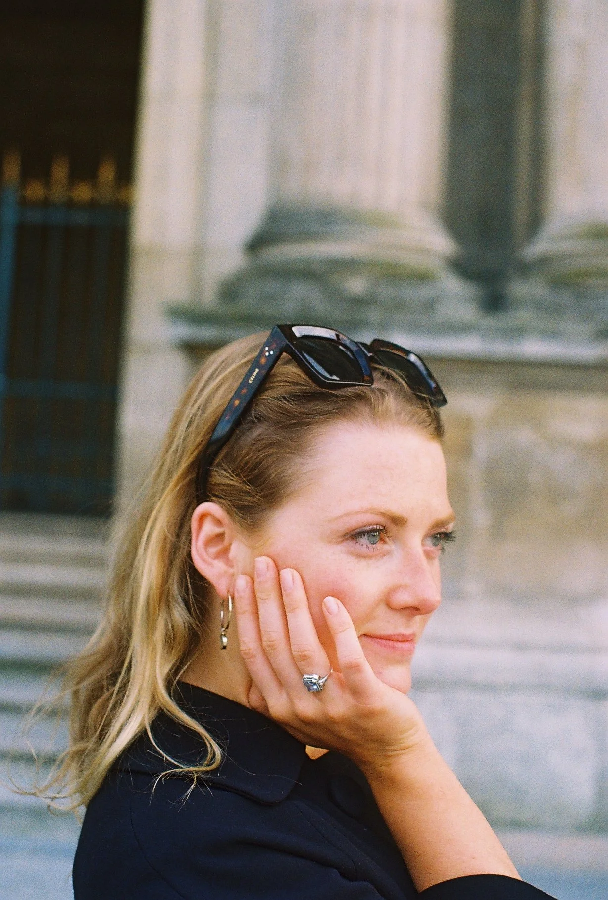 Femme avec lunettes de soleil sur la tête, portant une boucle d'oreille et une bague, posant près d'un bâtiment en pierre.