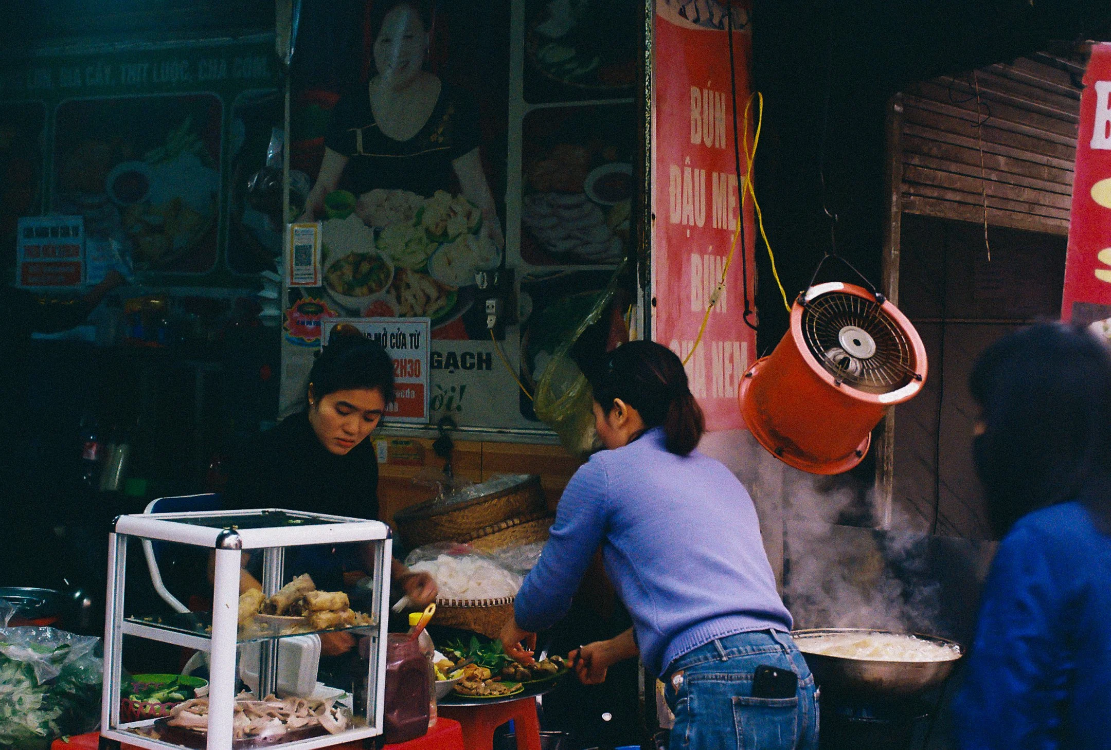 Une scène de marché de nuit où des femmes préparent et vendent des plats vietnamiens, avec de la vapeur s'échappant d'une grande casserole et une casserole orange suspendue.