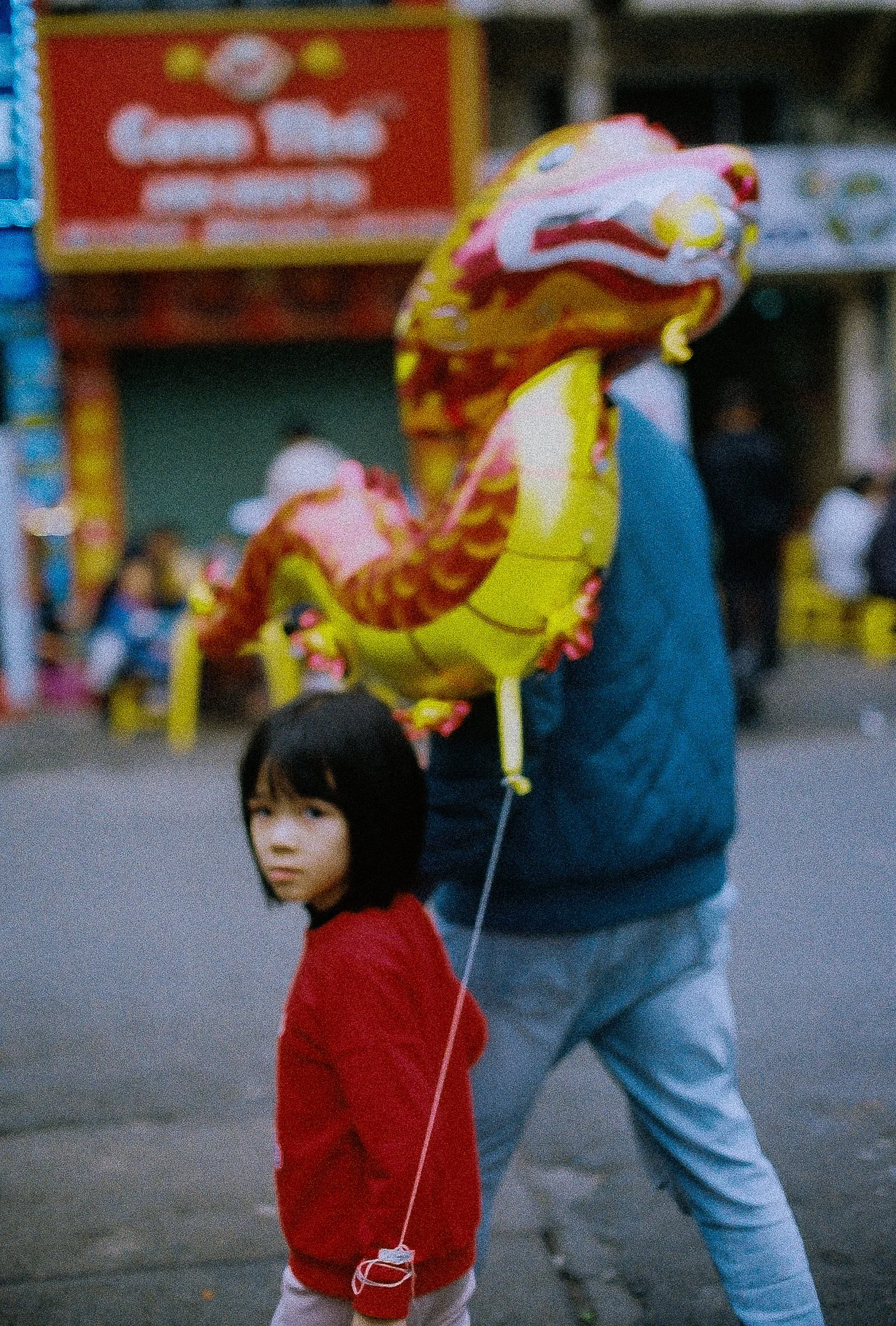 Une jeune fille avec un ballon en forme de dragon observe la caméra, tandis qu'un adulte porte un ballon en forme de dragon derrière elle, dans une rue animée.