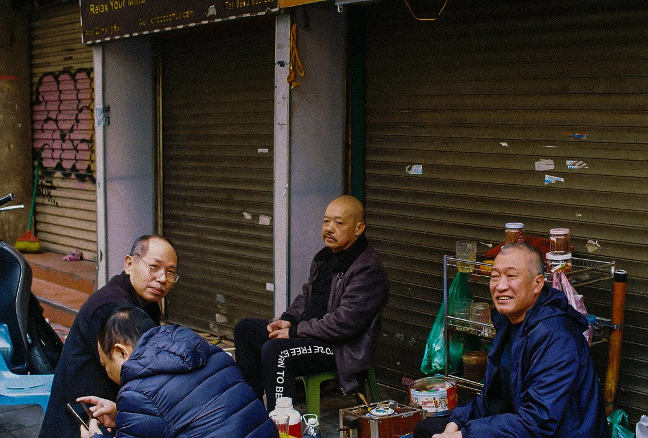 Quatre hommes tenant une conversation dans une rue, certains assis sur des petits tabourets, avec des étals de nourriture et des roulettes de ferraille en arrière-plan.