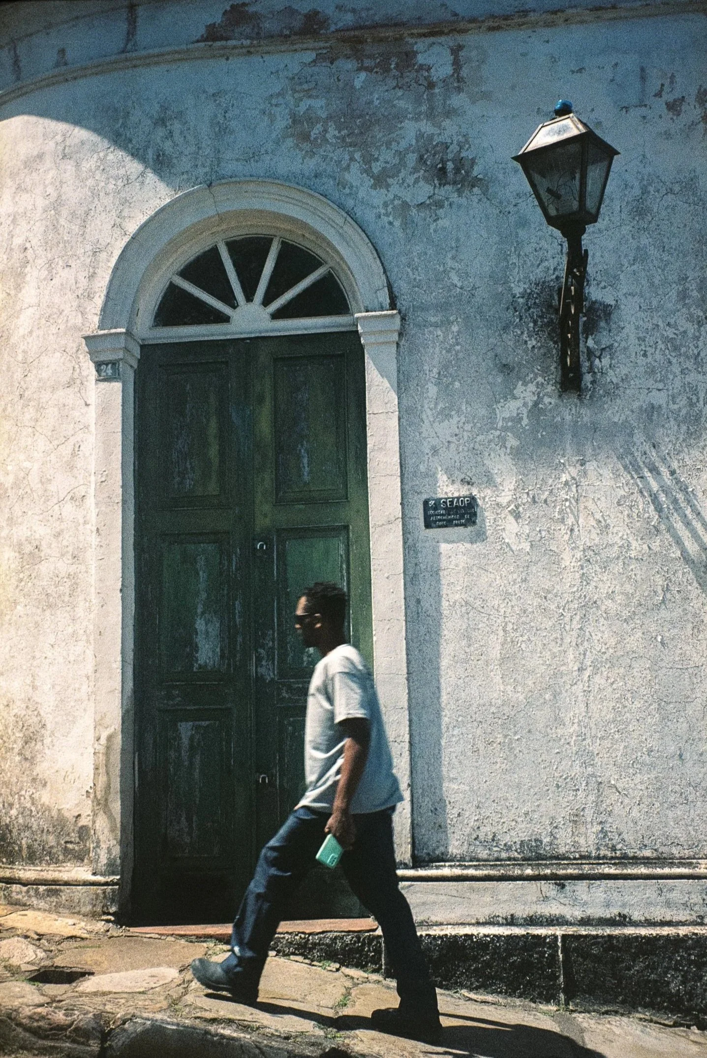 Un homme marchant devant une vieille façade blanche avec une porte en bois vert et une lanterne en métal noir fixé au mur.