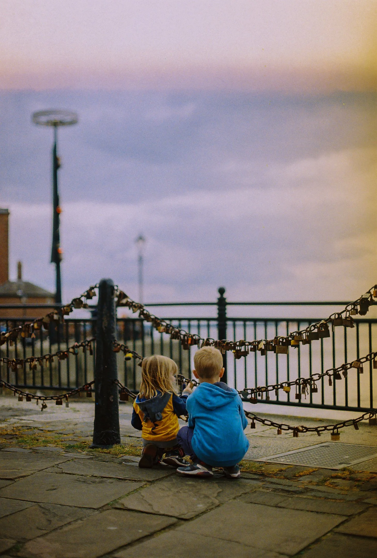 Deux enfants assis sur le trottoir, regardant par-dessus une barrière en fer forgé avec des cadenas, sous un ciel au crépuscule.