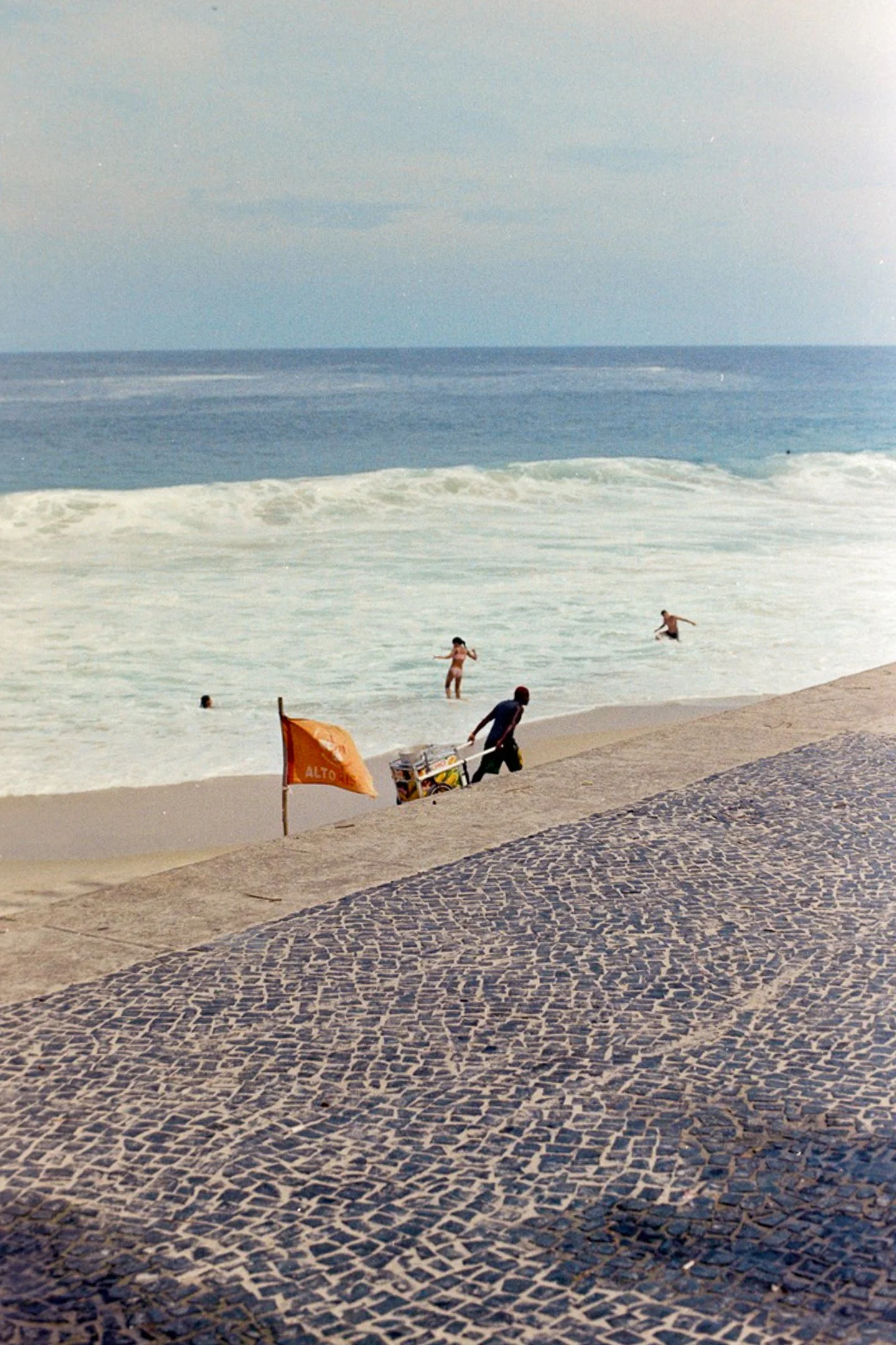 Personnes nageant dans la mer à la plage, avec un sauveteur et un drapeau orange sur la plage.