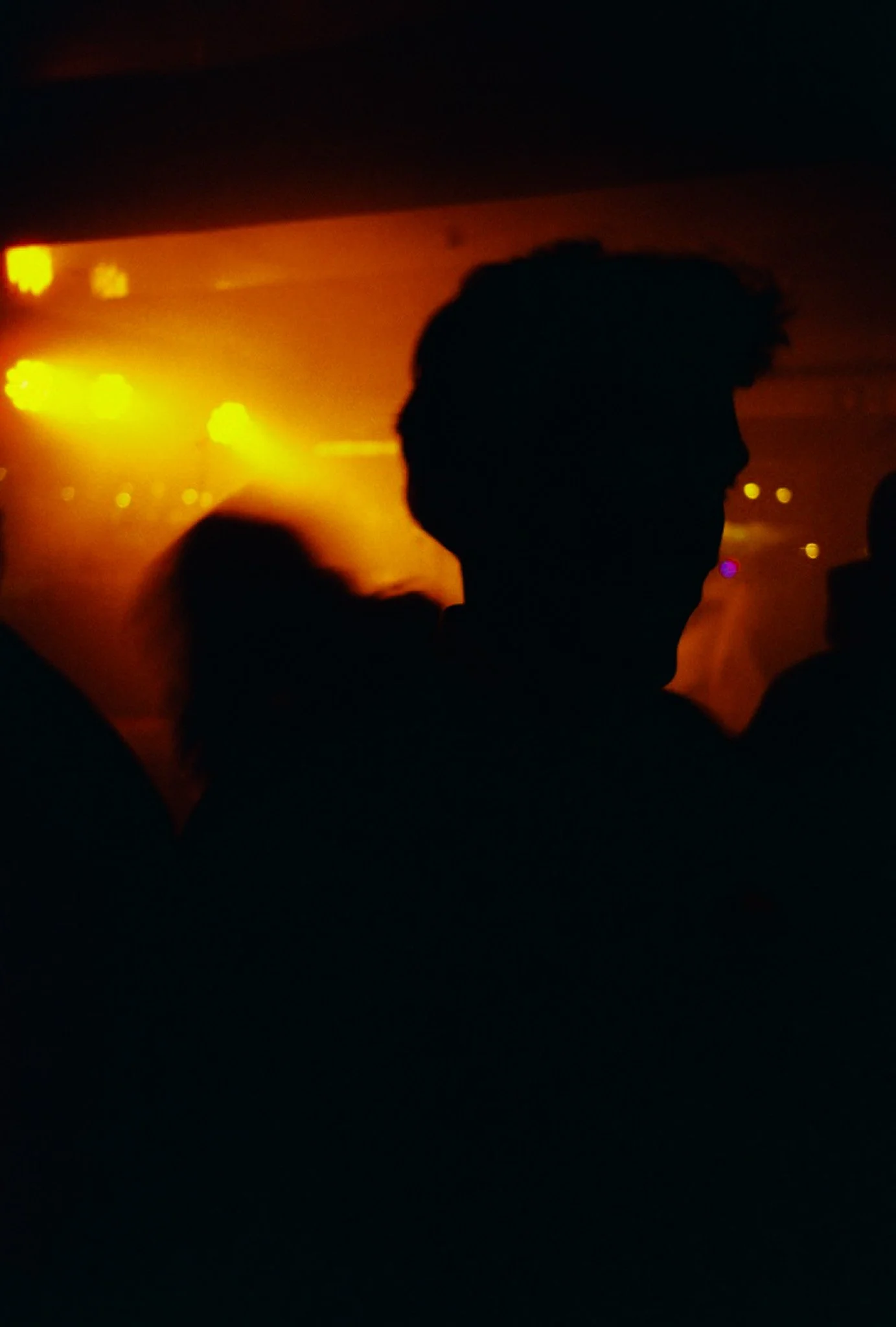 Silhouette d'une personne dans une ambiance sombre avec des lumières jaunes et oranges en arrière-plan, probablement lors d'une fête ou d'un concert.