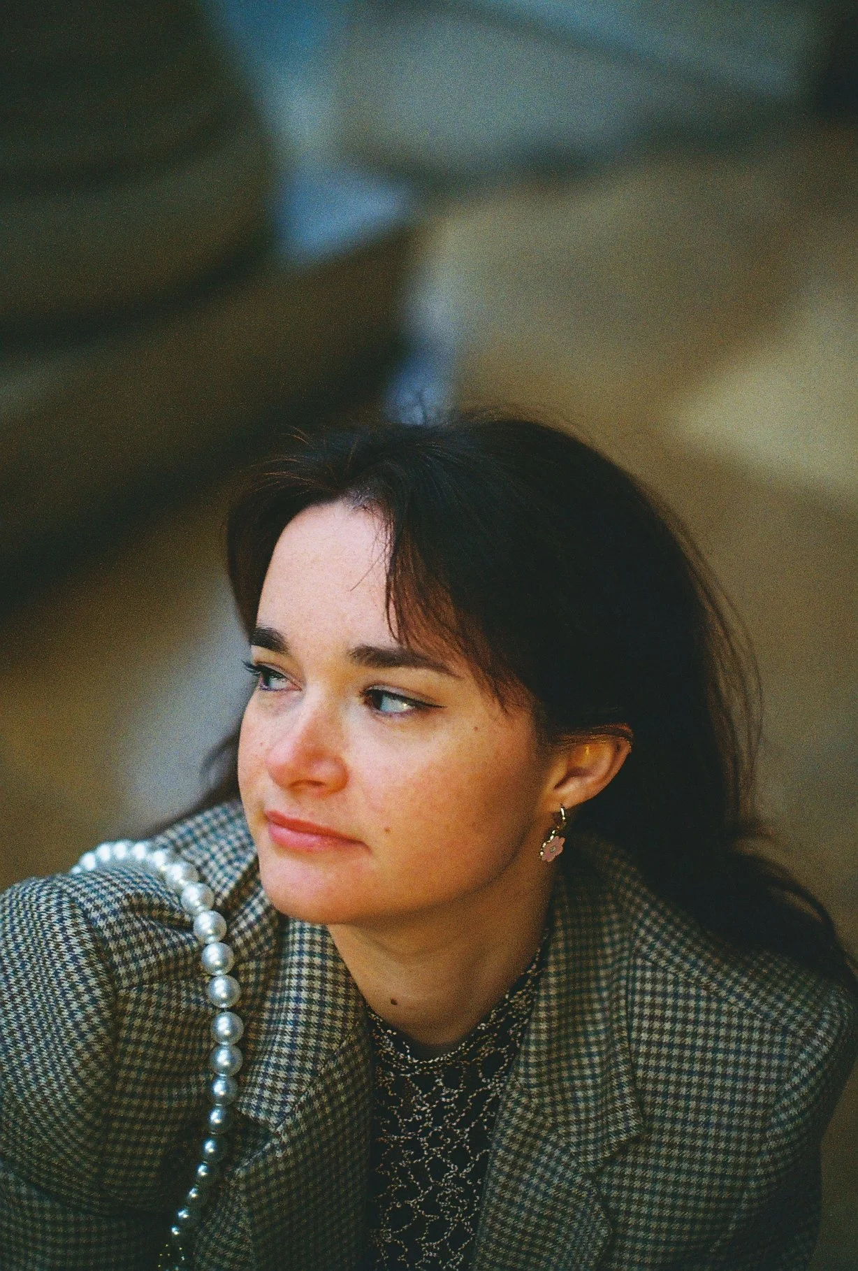 Jeune femme aux cheveux bruns foncés, regardant vers la gauche, portant un blazer à carreaux, un haut noir à motifs, des boucles d'oreilles roses et une chaîne de perles.