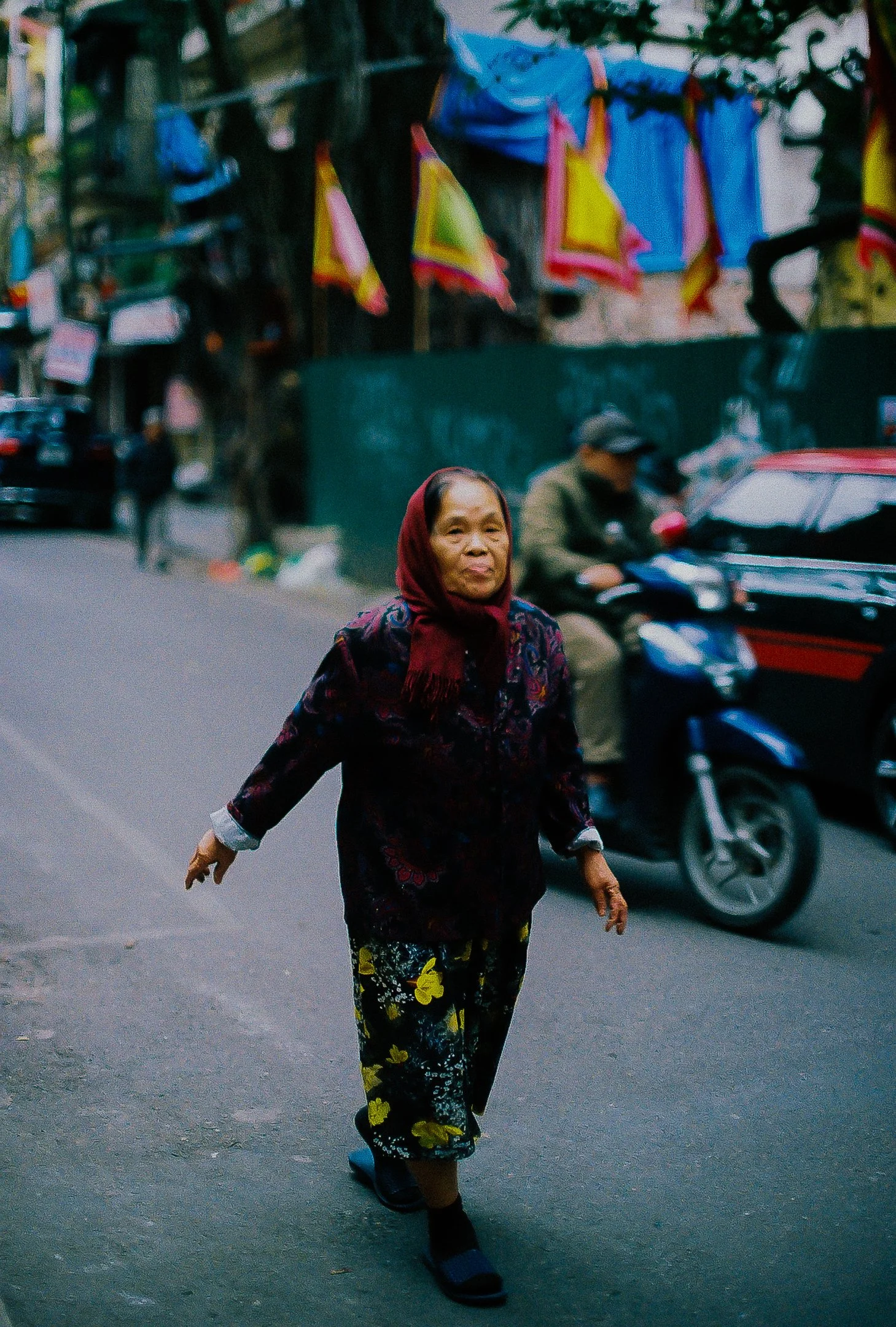 Une femme âgée marche sur une rue urbaine, portant un foulard rouge et des vêtements colorés, avec des voitures et des personnes en arrière-plan.