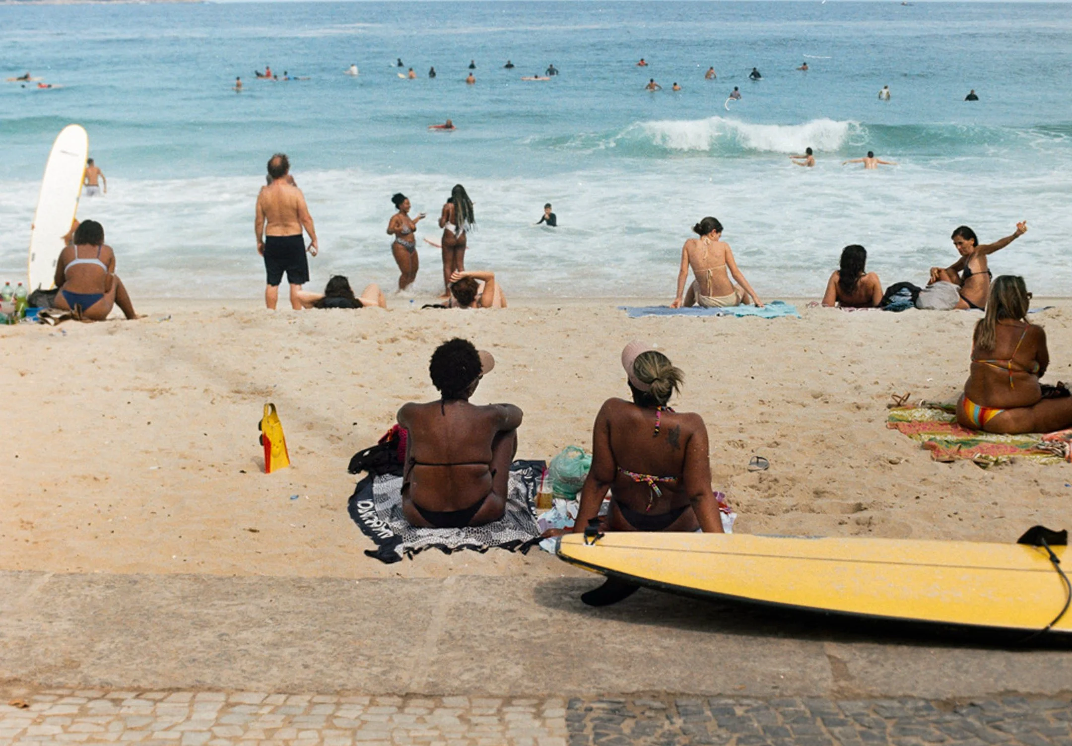 Plage avec plusieurs personnes, some seated on the sand, some in the water, some standing or lying down, avec une planche de surf sur le sable.