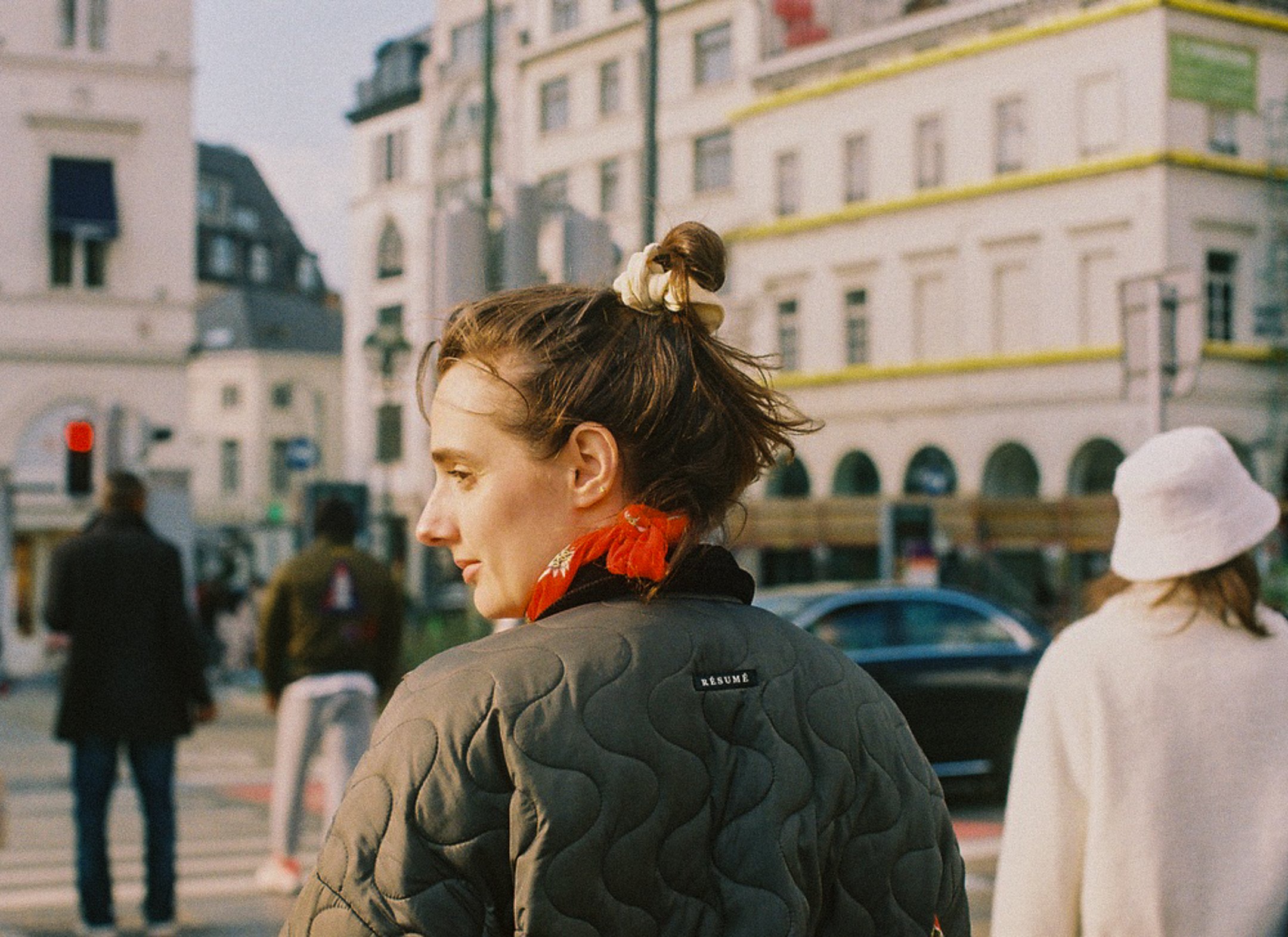 Une femme avec des cheveux bruns attachés en un chignon, portant une écharpe rouge et une veste molletonnée noire, regarde vers la gauche dans une rue urbaine, avec d'autres personnes et des bâtiments en arrière-plan.
