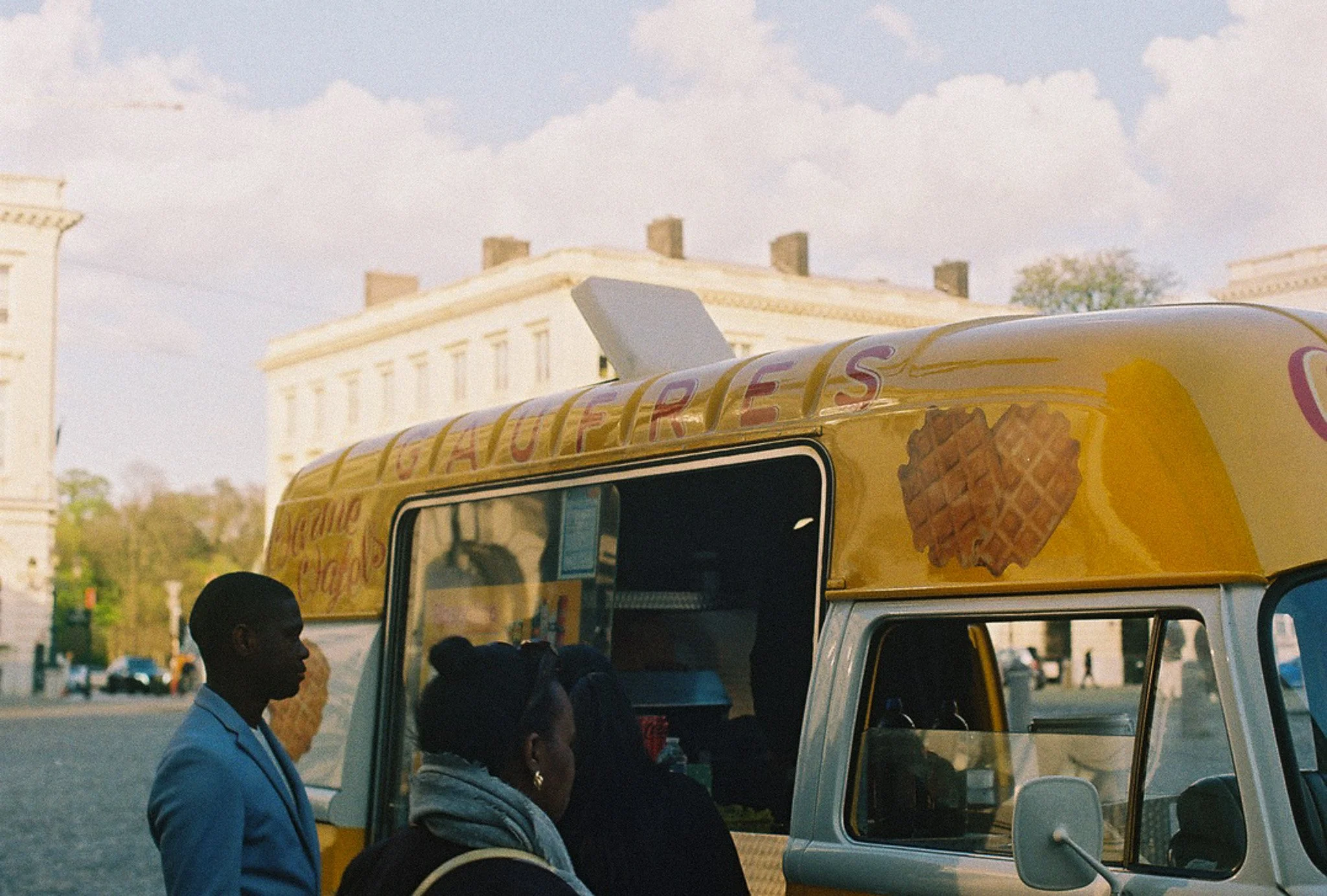 Un groupe de personnes autour d'une camionnette à glace dans un espace public en plein air, avec un bâtiment historique en arrière-plan, par une journée ensoleillée.