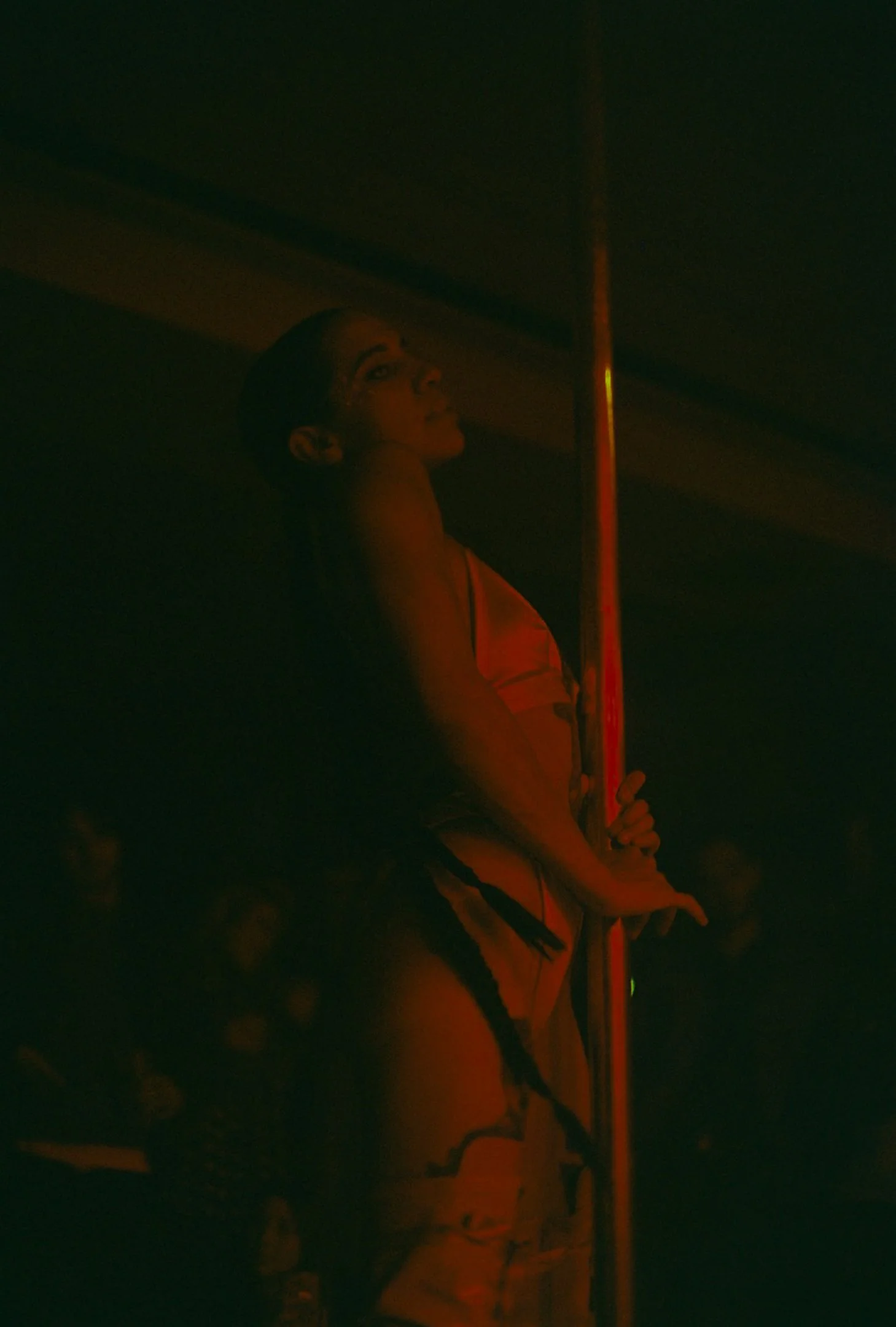 Femme dans un club de strip-tease, portant une tenue colorée, tenant un poteau de pole dance, ambiance sombre et éclairée par une lumière rouge
