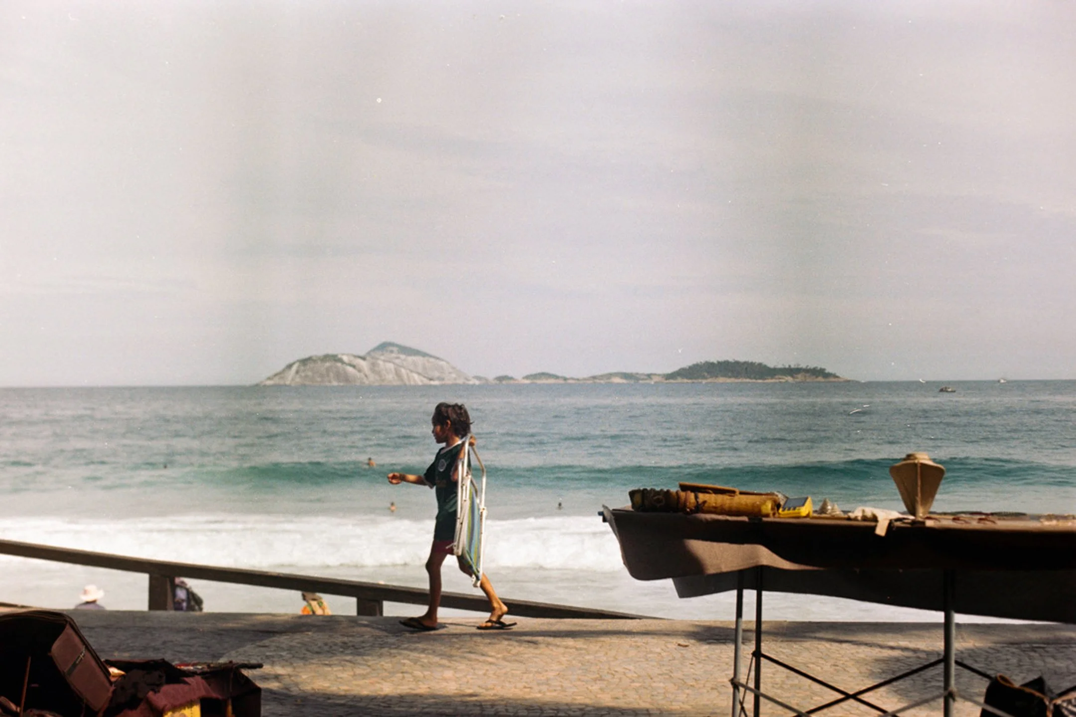 Une personne marchant sur une promenade en bord de mer avec des îles en arrière-plan, un bateau à l'avant-droite et des affaires de plage sur le sol.
