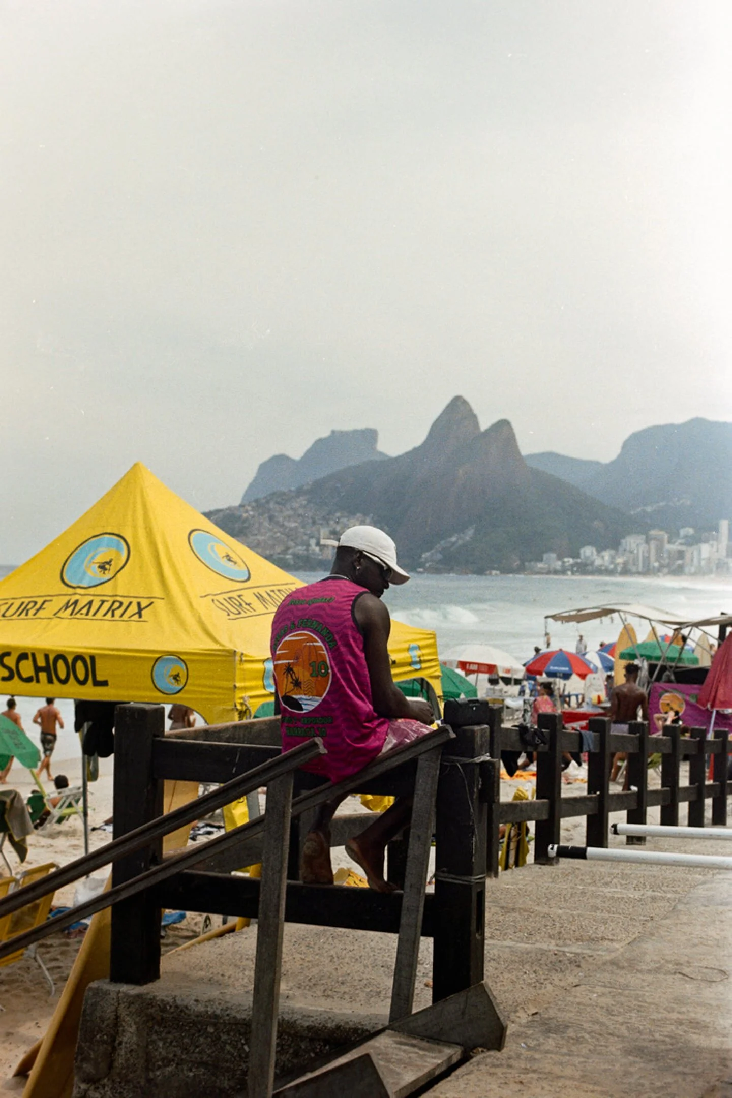 Person assise sur une plateforme en bois au bord de la plage, portant un t-shirt rose, une casquette blanche, avec des parasols colorés et la mer en arrière-plan, ainsi que des montagnes et des bâtiments au loin.