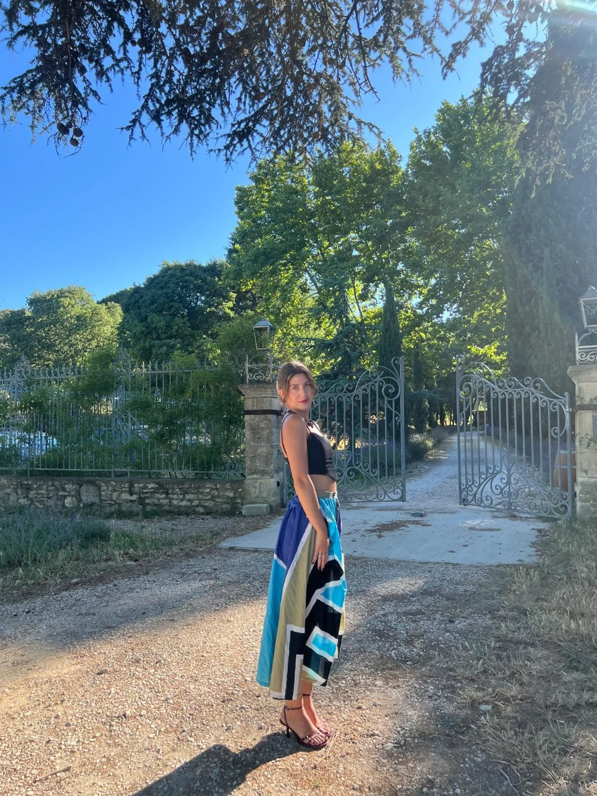 Jeune femme en robe longue colorée, debout devant une grande porte en fer forgé, entourée de verdure et d'arbres sous un ciel bleu.