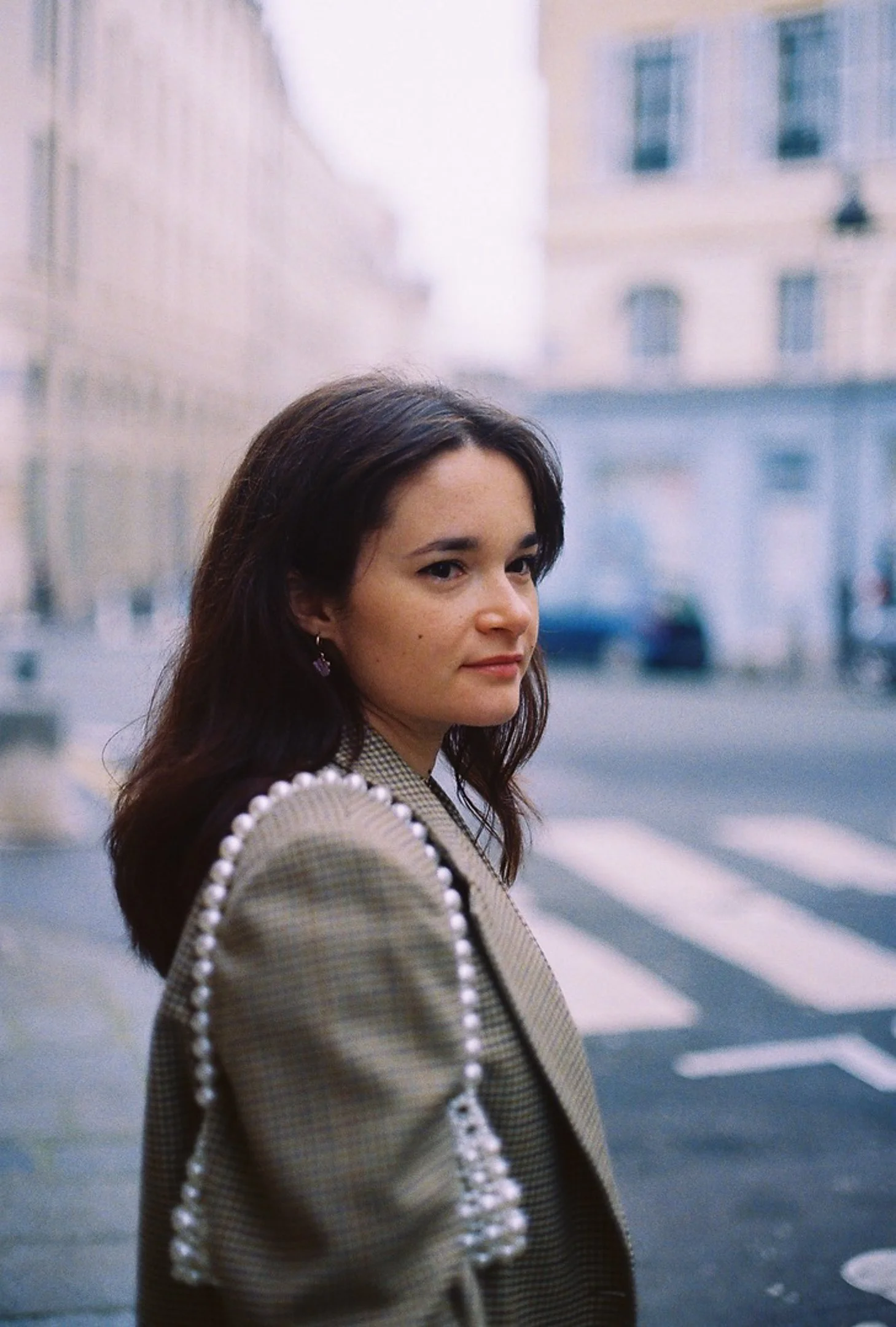 Jeune femme avec cheveux bruns, vêtue d'un blazer à épaule perlée, en ville.