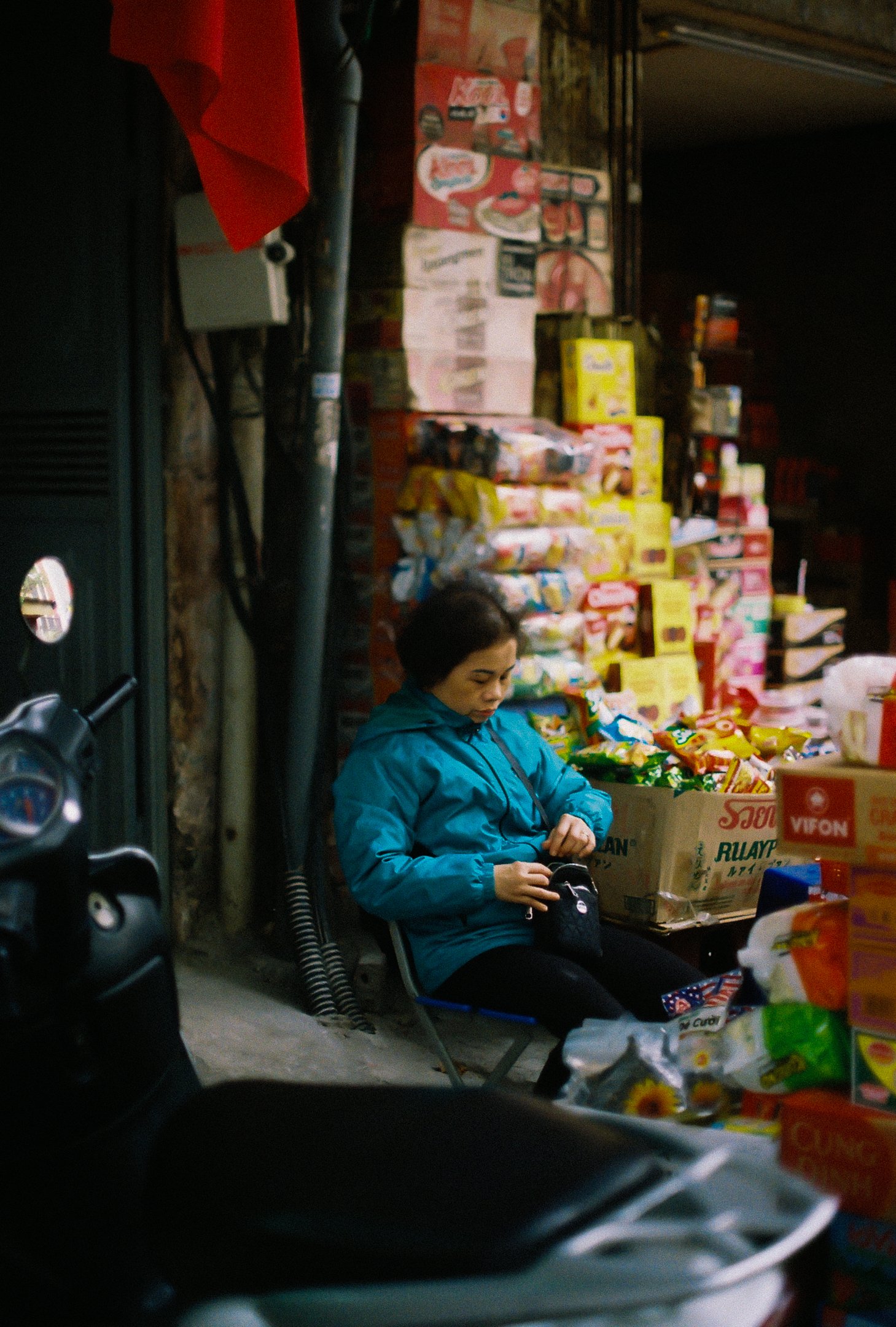 Une femme assise sur une chaise à côté d'un stand de marchandises, portant une veste bleue, à l'extérieur d'une boutique ou d'un marché, entourée de boîtes de produits divers.