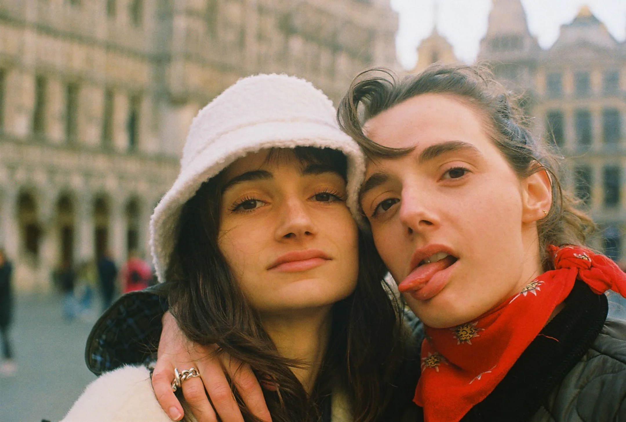 Deux jeunes personnes posent ensemble dans une ville, l'une porte un chapeau blanc et l'autre un bandana rouge, exprimant une attitude décontractée et amusante.