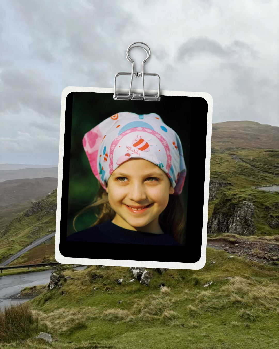 Une photo d'une jeune fille souriante portant un chapeau coloré, suspendue par un clip dans un paysage de collines verdoyantes et ciel nuageux.
