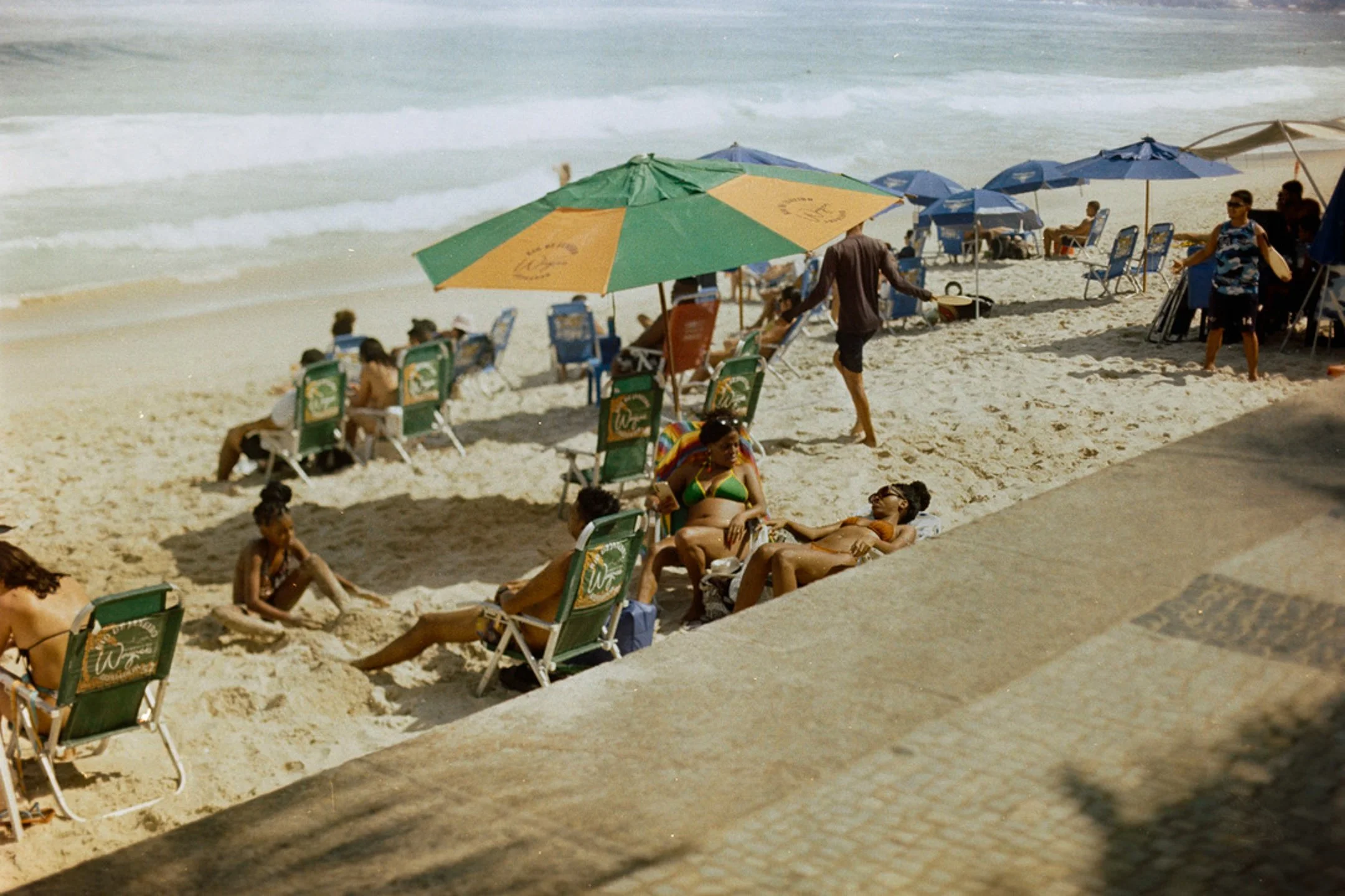 Plage de sable avec des parasols, des chaises longues et des personnes qui se détendent ou jouent dans le sable, avec la mer en arrière-plan.