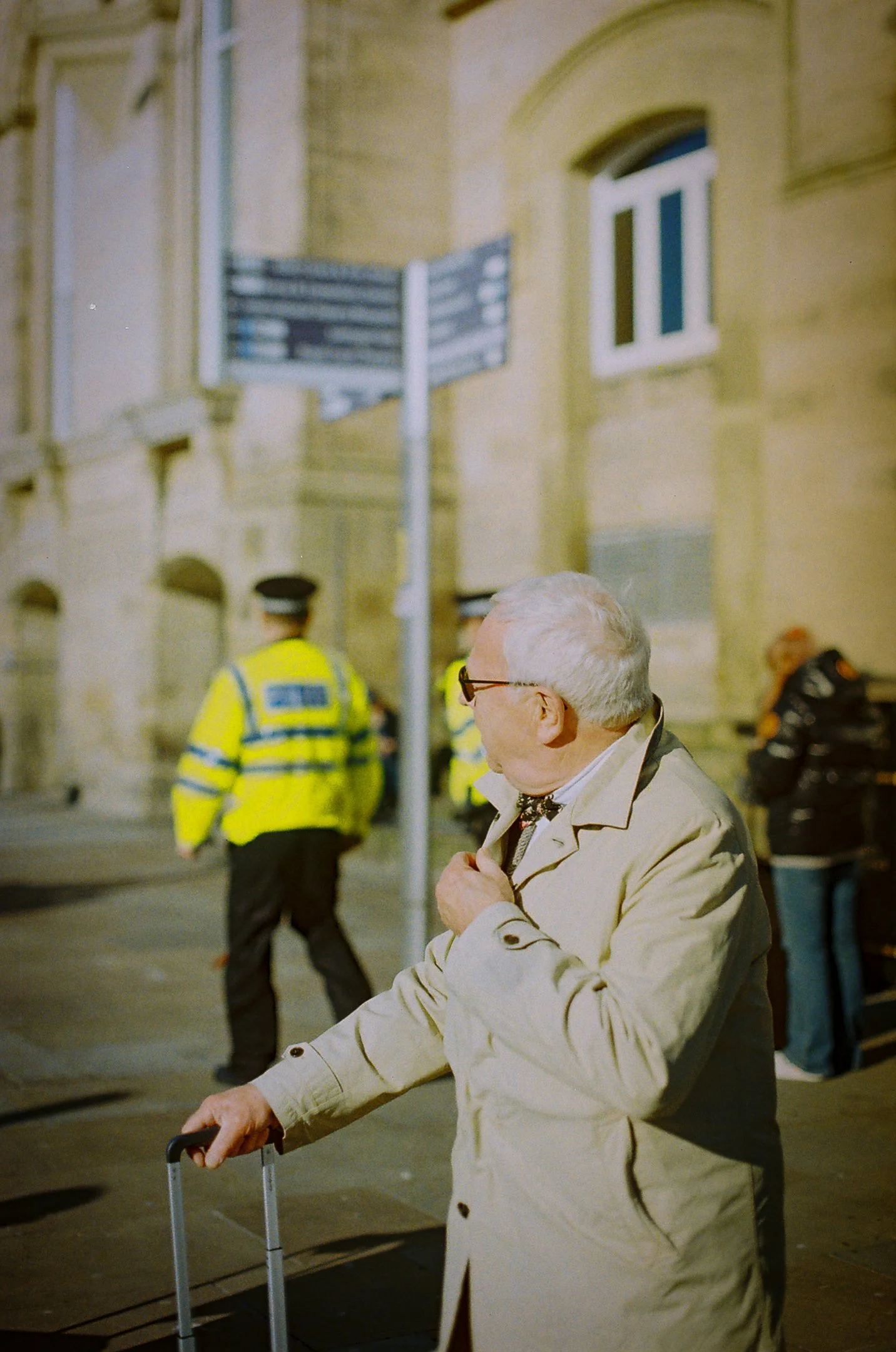 Un homme âgé en trench-coat clair et lunettes, tenant une valise à roulettes, regarde vers la droite dans une rue pavée, avec quelques policiers en vestes jaunes fluorescentes en arrière-plan et d’autres personnes dans la scène.