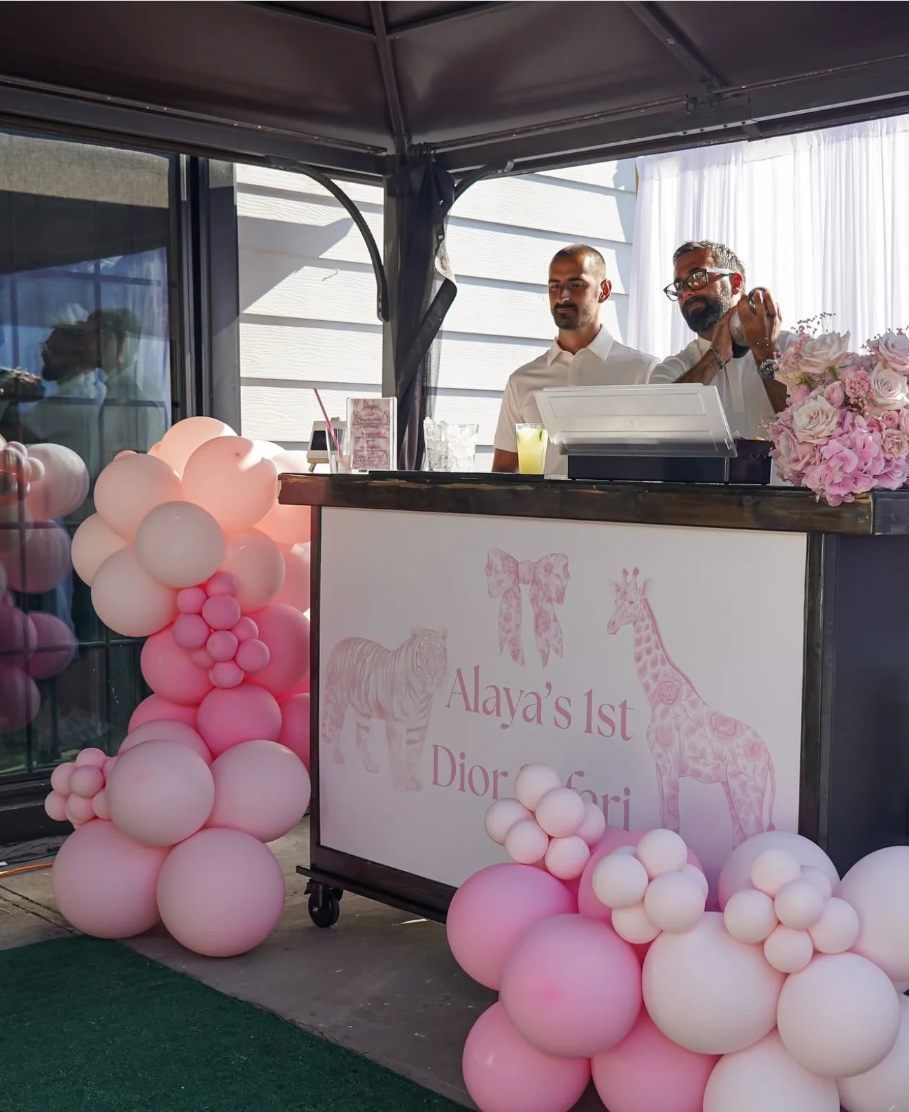 A decorated birthday party booth with pink and white balloons, a sign that reads 'Alaya's 1st Dior Safari' featuring illustrations of a tiger, a bow, and a giraffe, with two men behind the counter.