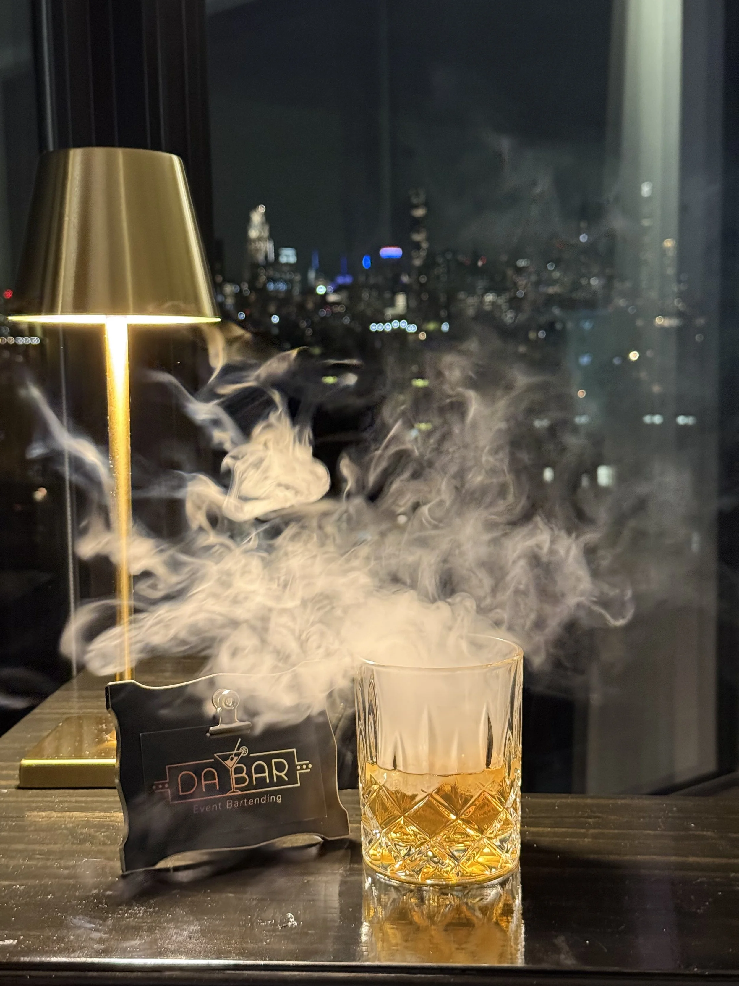 A glass containing an amber-colored drink with smoke rising from it, placed on a dark table near a window with a city skyline in the background. There is a small black sign with the text 'DA BAR Event Bartending' and a gold lamp to the left.