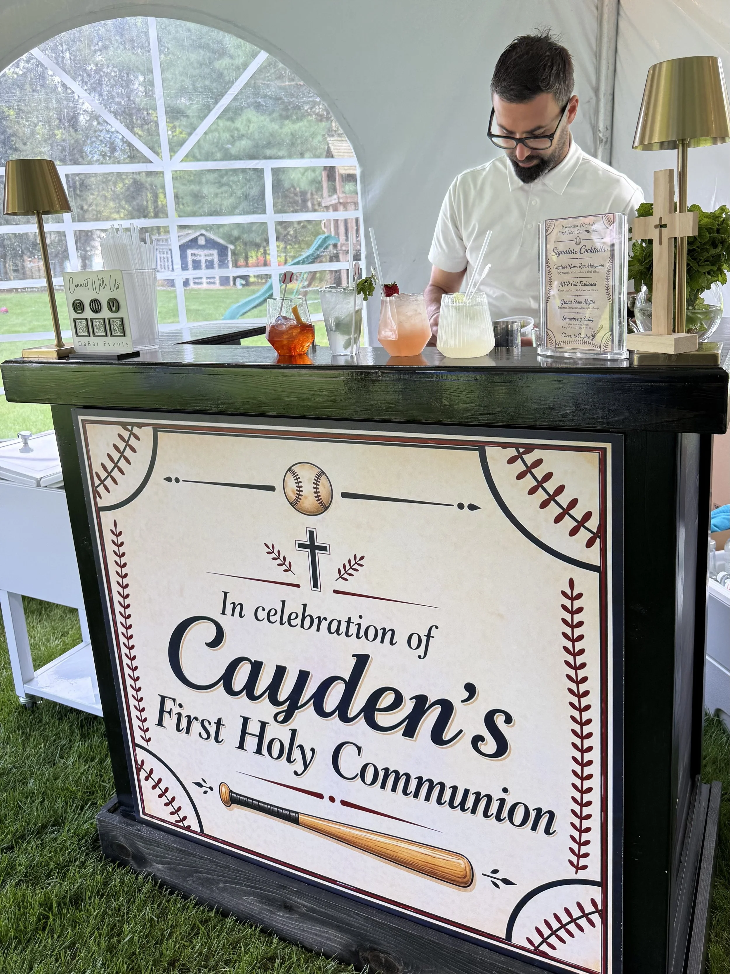 A decorated event sign celebrating a first holy communion, featuring baseball-themed design elements such as a baseball and a bat, and a list of signature cocktails in an outdoor setting with grass, trees, and a playground in the background.