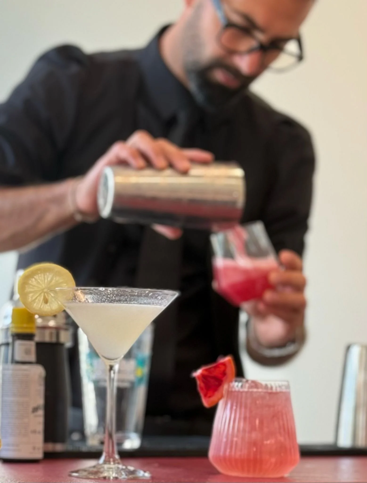 A bartender making a cocktail at a bar, with two completed drinks in the foreground: a pale yellow drink with a lemon slice garnish and a pinkish drink with a grapefruit wedge garnish.