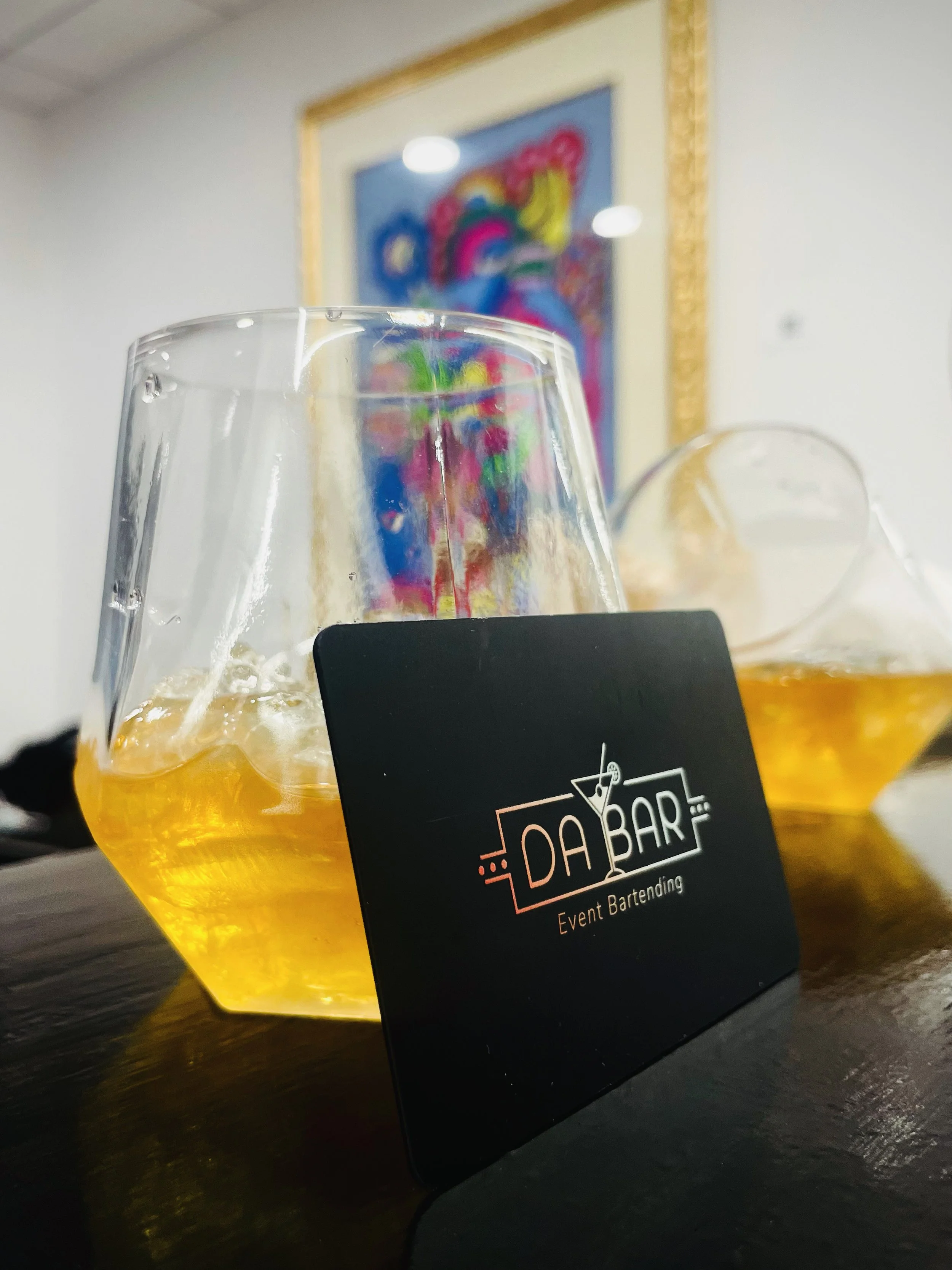 Two glasses of yellow-colored beverage with ice, placed on a dark table, with a black card reading "El Dabar Event Bartending" leaning against one of the glasses. A colorful framed painting is visible on the wall in the background.