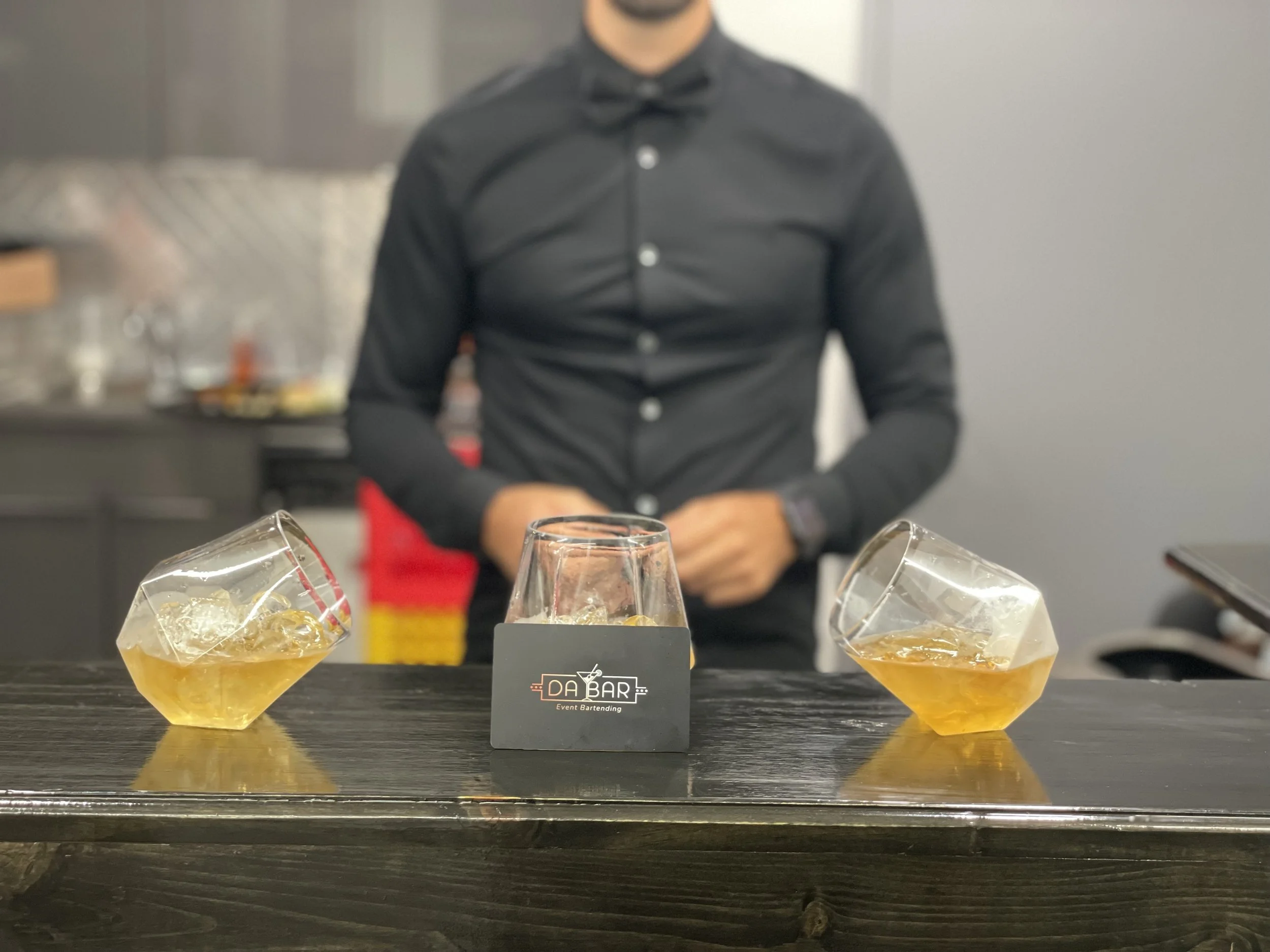 Three glasses of whiskey on a bar counter, two tilted and one upright, with a bartender in the background wearing a black shirt.