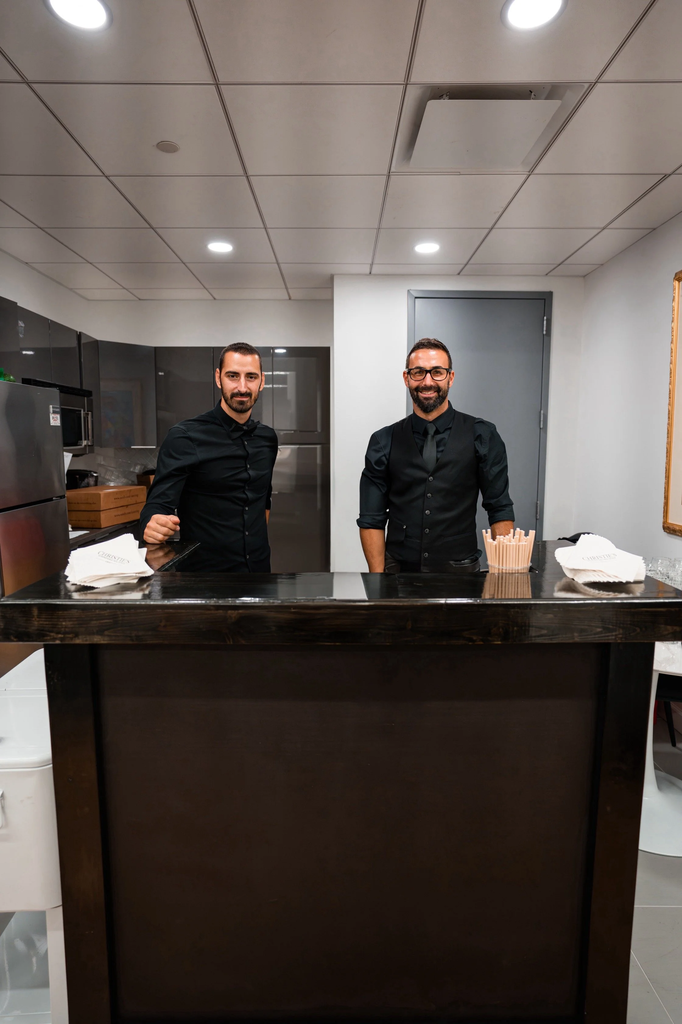 Two men standing behind a dark wooden bar counter in a modern kitchen, both dressed in black shirts and smiling for the photo.