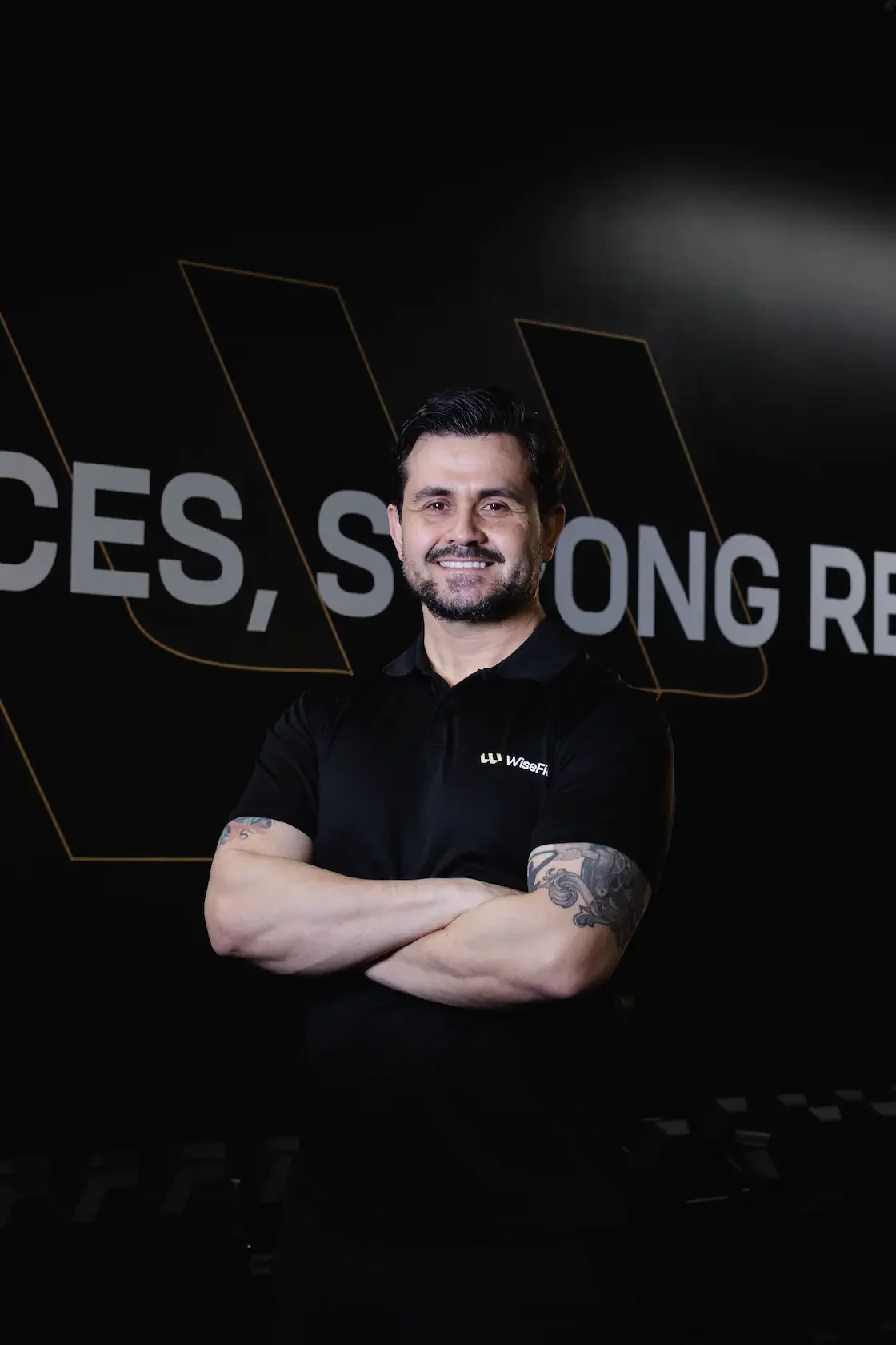 A man with dark hair and a beard, smiling with arms crossed, wearing a black WiseFin polo shirt, standing in front of a black background with partial text and geometric designs.