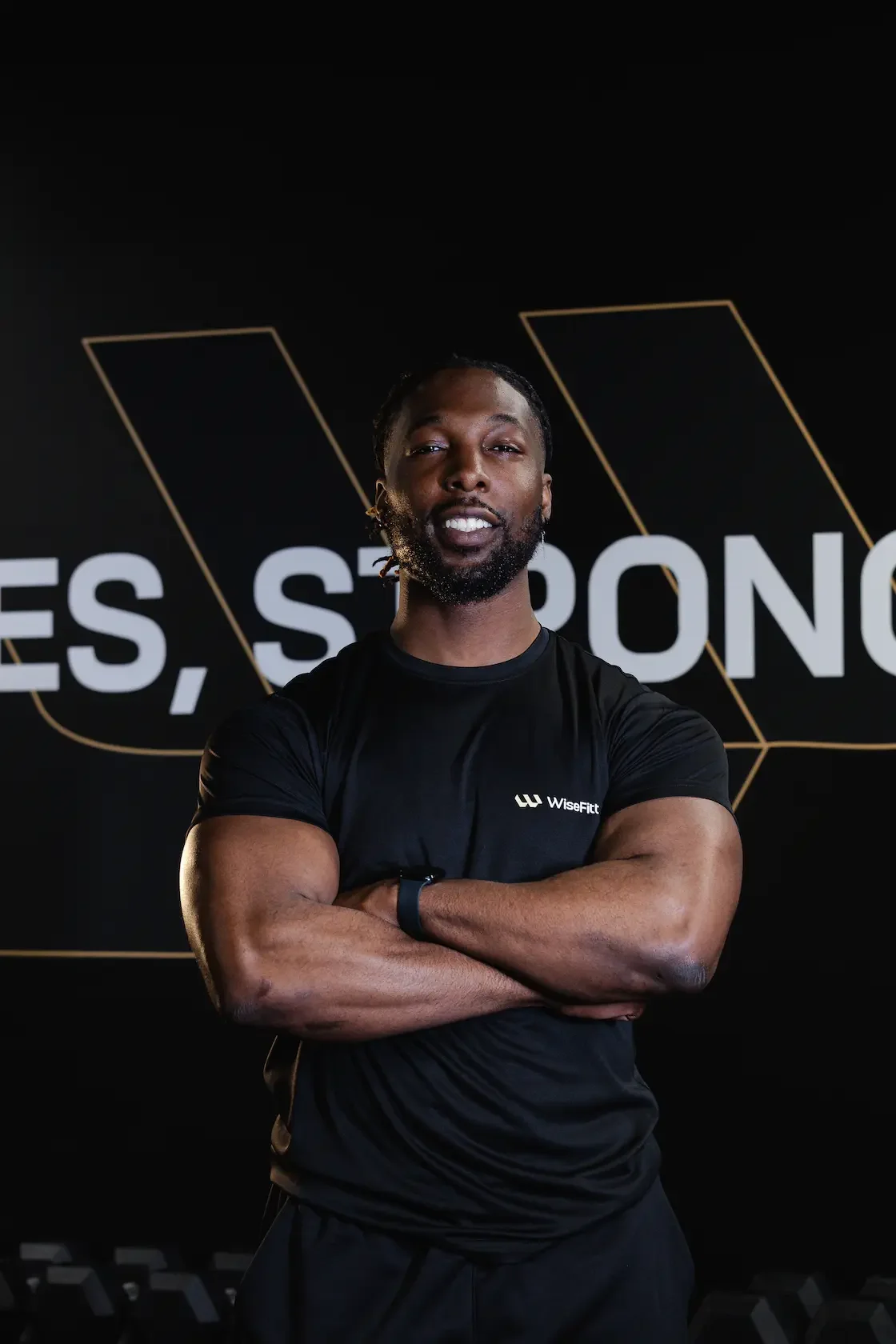 A muscular man with crossed arms wearing a black WiseFit t-shirt standing in front of a black background with white text and geometric designs.