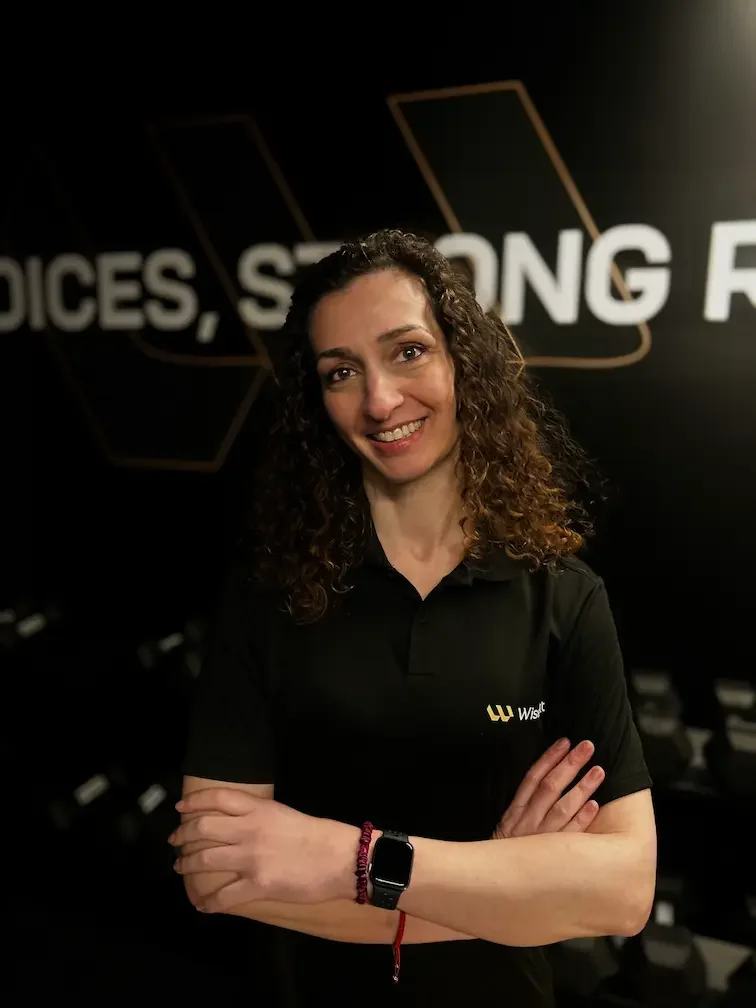 A woman with curly brown hair smiling, standing with arms crossed in front of a dark background with partially visible text.