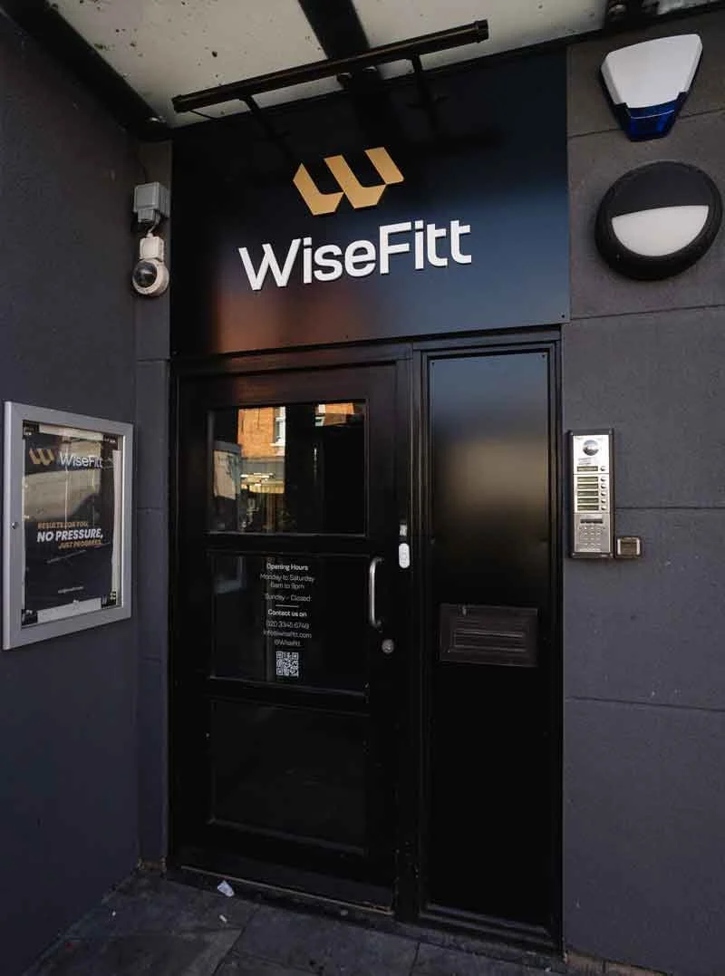 Black entrance door for WiseFitt gym with sign above and a small window. Exterior wall in dark gray with security camera and orange brick building reflection visible.