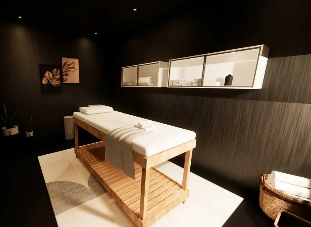 A massage therapy room with a massage table, pillows, and white towels, dark wood walls, decorative artwork, shelves with amenities, and a wicker basket.