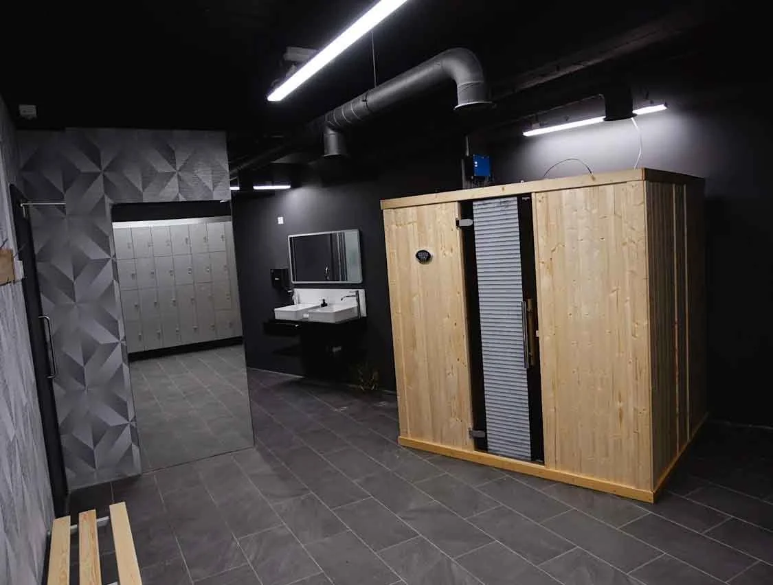 Indoor gym locker room area with lockers, a mirror with sink, and a sauna or steam room made of wood.