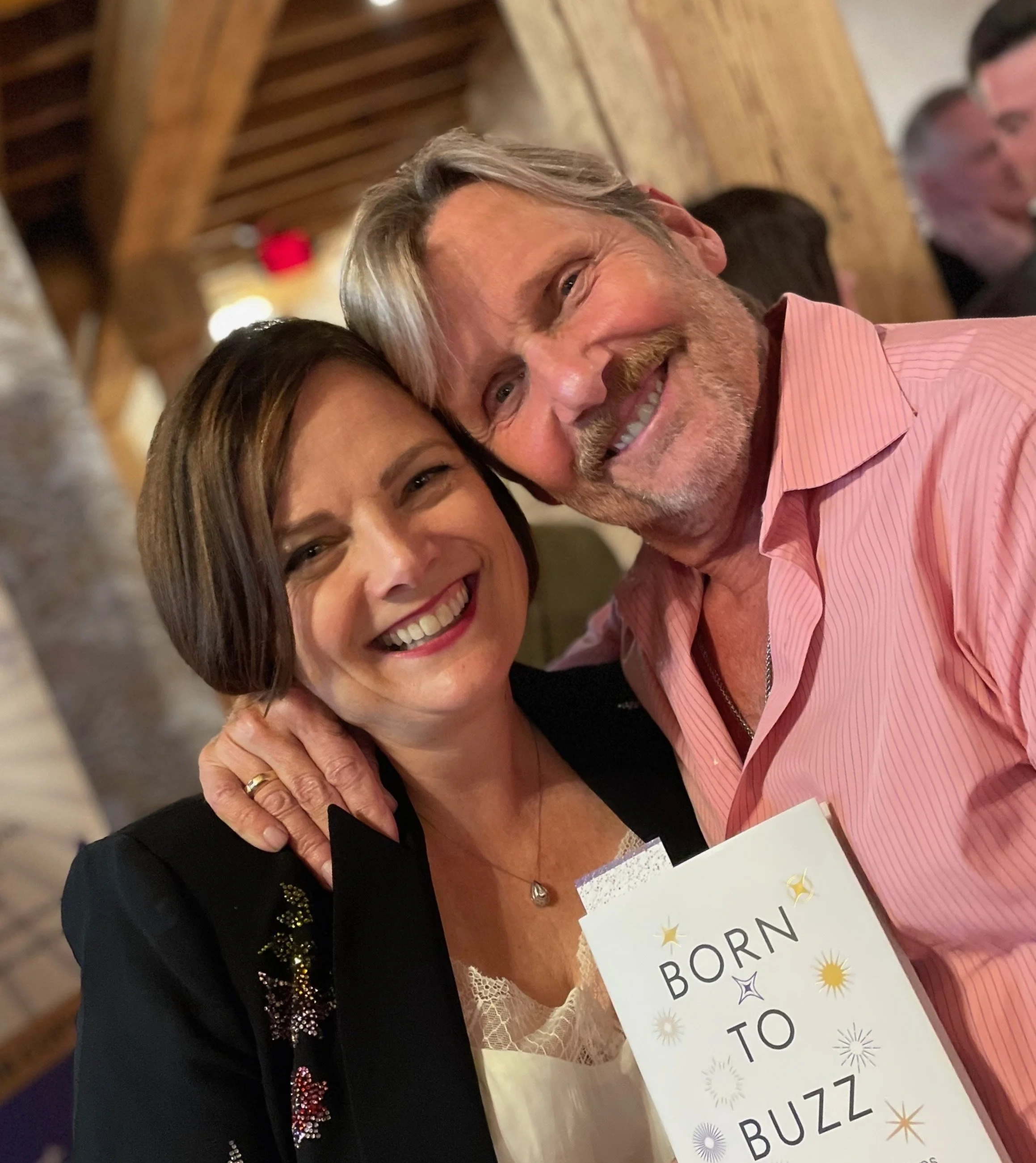 A smiling man and woman taking a selfie, with the woman holding a book titled 'Born to Buzz'.