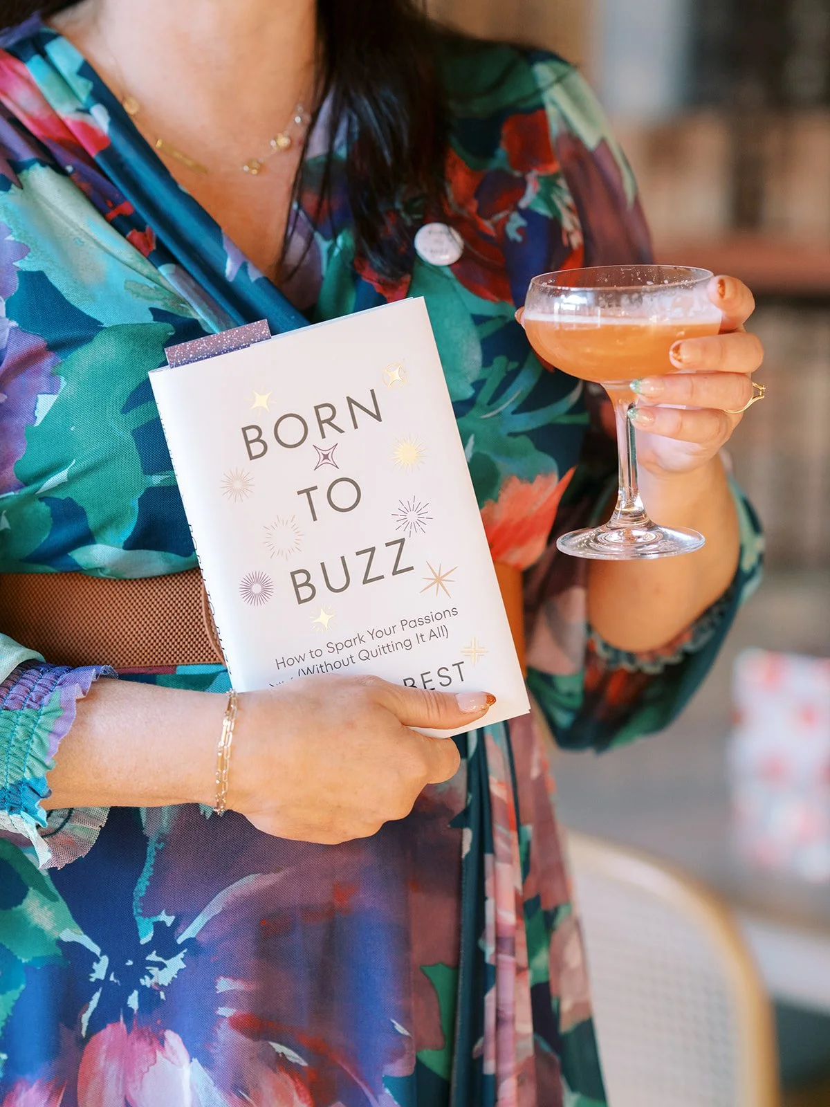 A woman holding a book titled 'Born to Buzz' and a pink cocktail, wearing a colorful floral dress with jewelry.