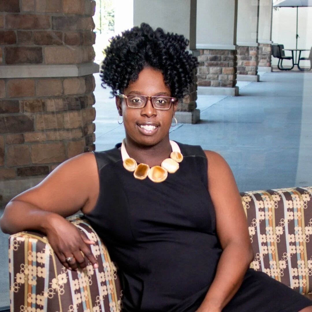 A woman with natural curly hair, glasses, and hoop earrings, wearing a sleeveless black top and a shell necklace, seated on a patterned bench in a modern outdoor plaza.