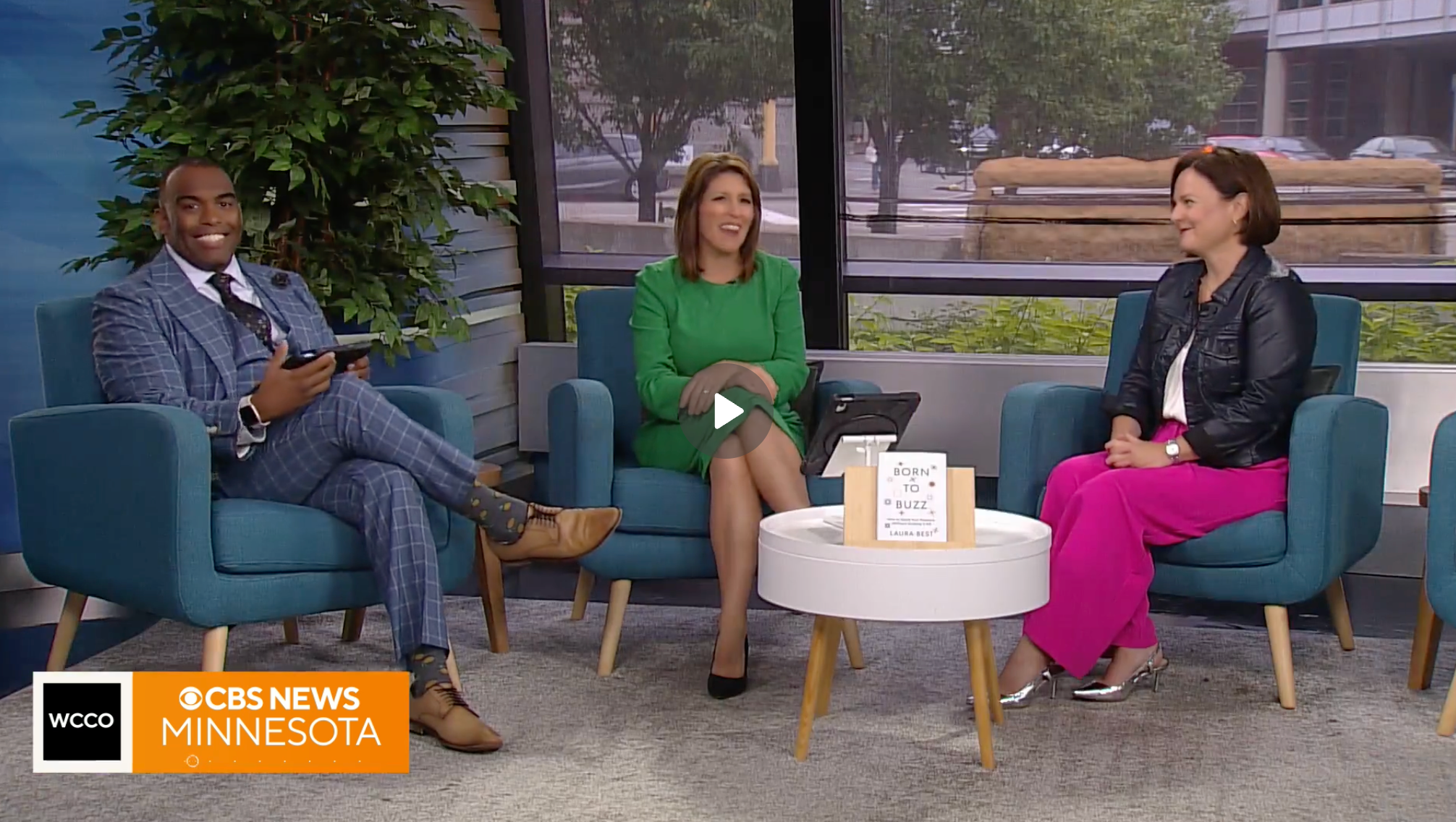 Three news anchors seated on blue armchairs in a TV studio, with large windows showing trees and a street outside. The woman in the center wears a green dress, the woman on the right wears a black leather jacket and pink pants, and the man on the left wears a blue checkered suit. A white table with a sign reading 'Born to Buzz' is in front of them, and a CBS News Minnesota logo is at the bottom left corner.