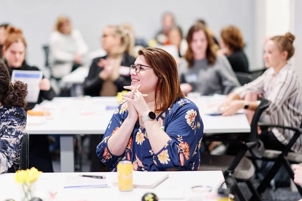 A woman with glasses and a floral blue blouse smiling and clapping at a conference or meeting with other women in the background.