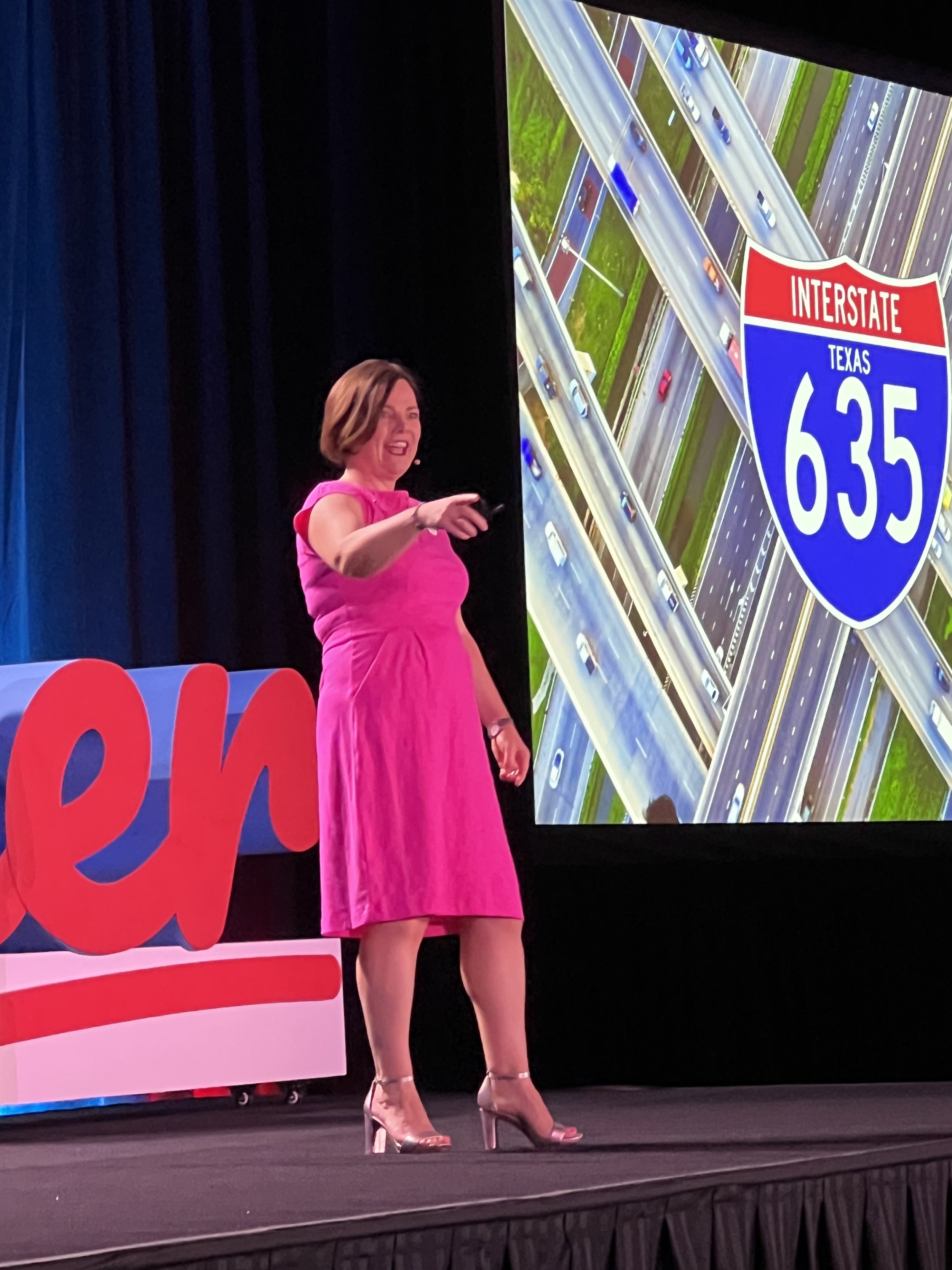 Woman in a pink dress giving a presentation on stage with a large screen showing a highway sign for Interstate 635 in Texas and the word 're' in red letters on stage.