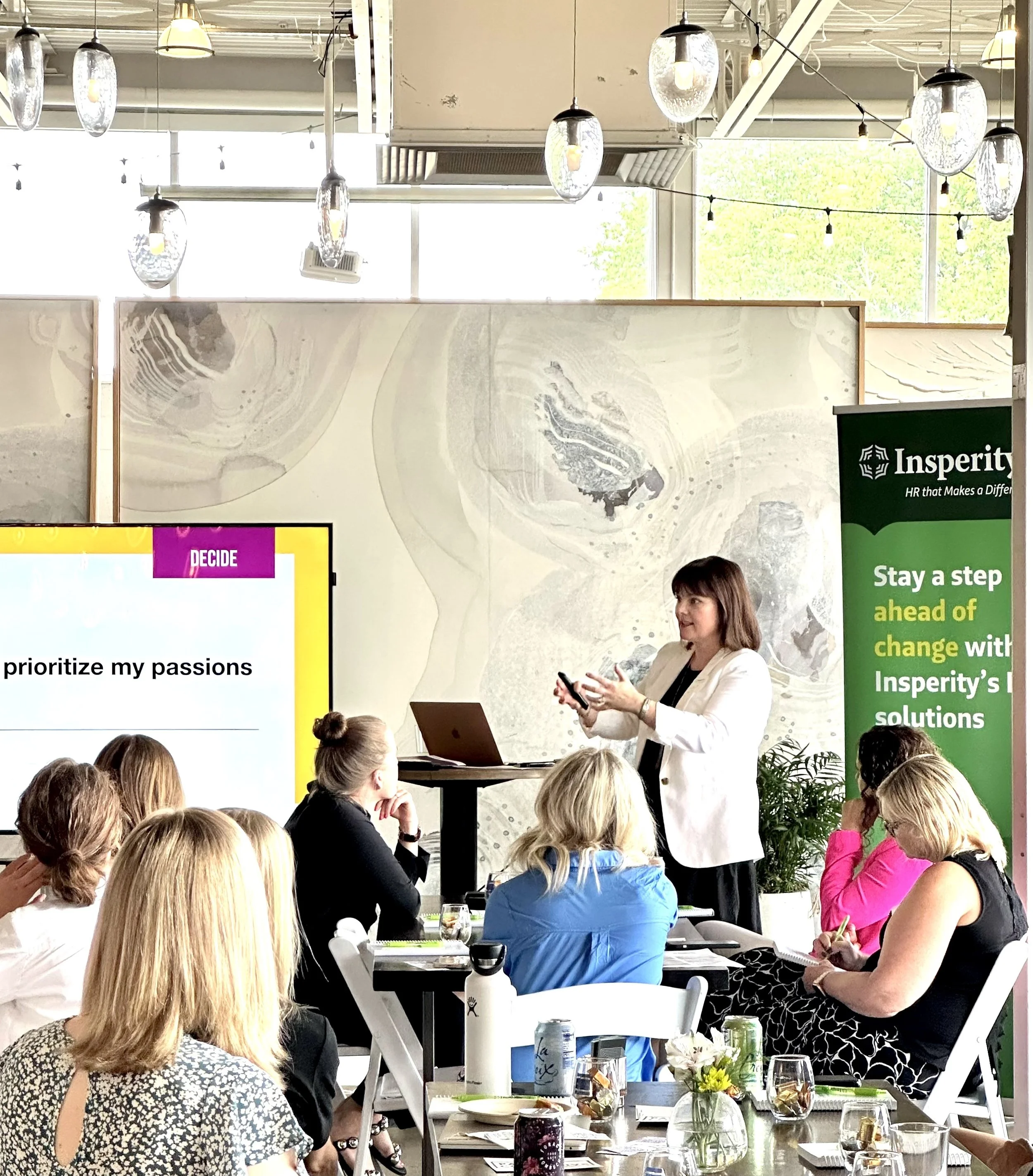 A woman in a white blazer presenting at a conference, standing in front of a large yellow and pink slide that says 'DECIDE' and 'prioritize my passions'. Several attendees, mostly women, are seated at tables, listening to her. There's a large banner on the right promoting Insperity with green and white colors.