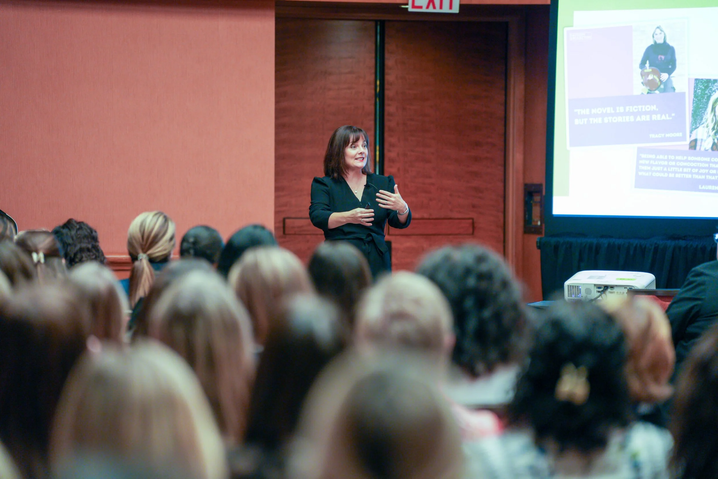 A woman in a black outfit giving a presentation to a seated audience in a conference room. A large screen displays slides on her right.