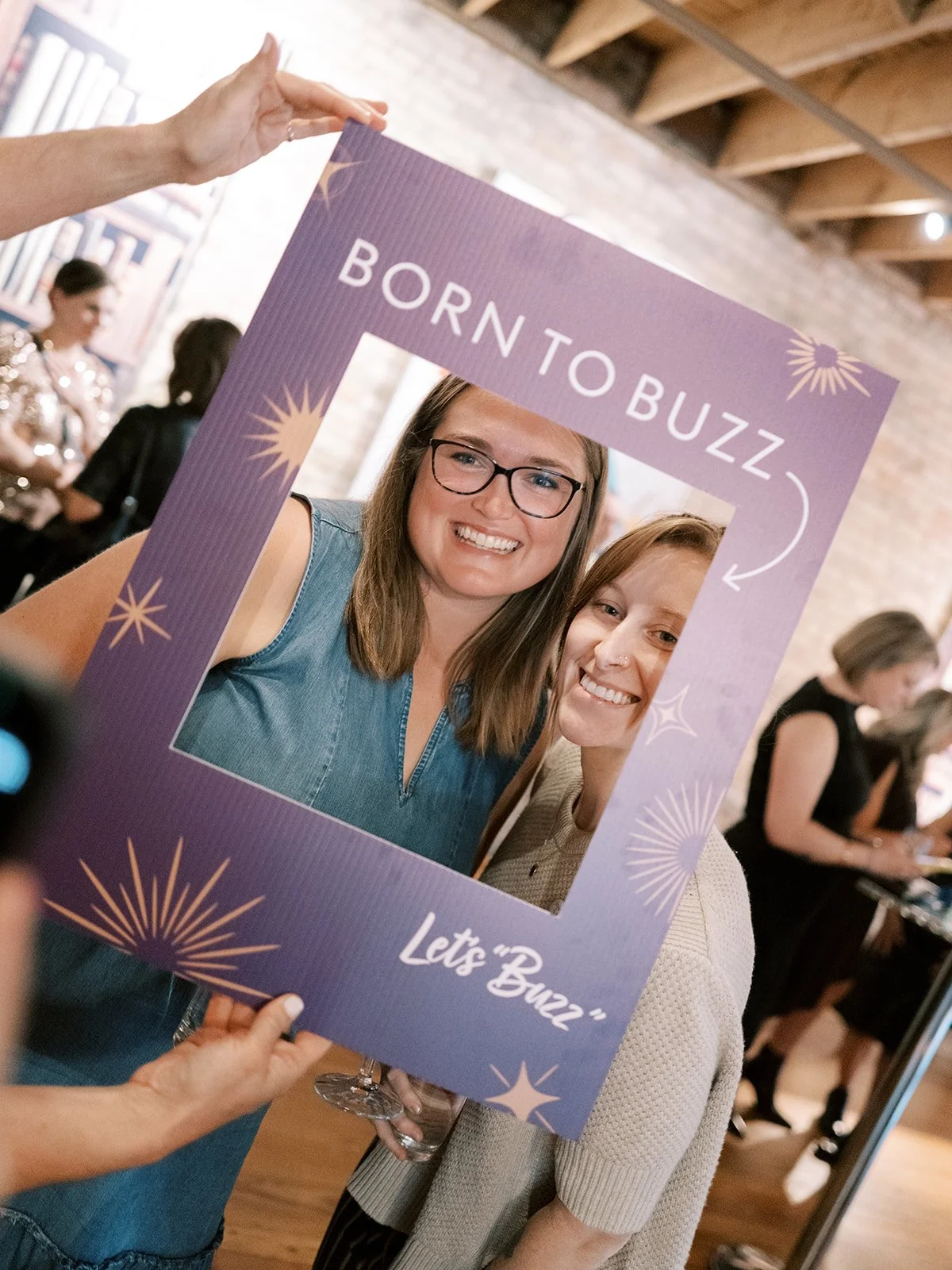 Two women smiling behind a large photo frame prop that reads 'Born to Buzz' and 'Let's Buzz' at an indoor event.